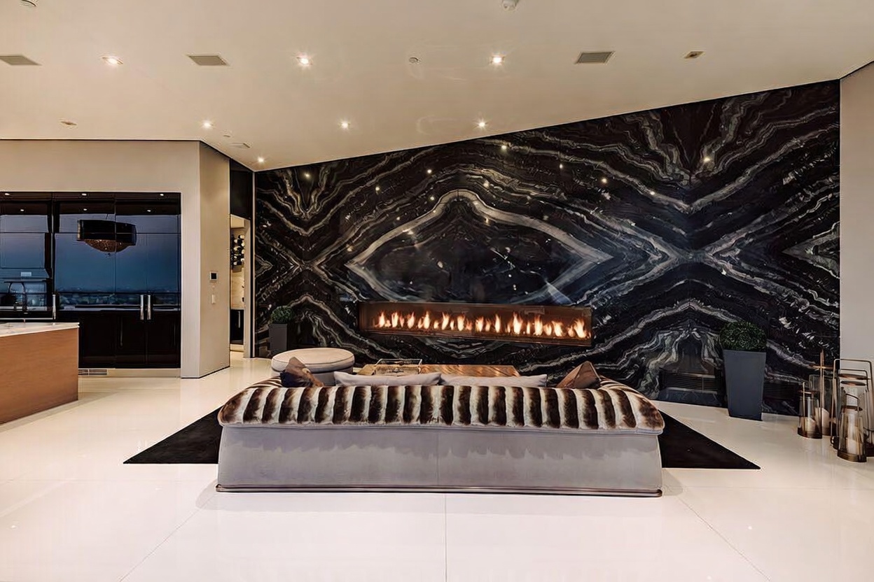 This luxurious living room features a striking, book-matched black marble accent wall with a sleek, linear fireplace as its centerpiece. A plush, textured sofa sits on a dark rug against the polished white flooring, creating a sophisticated and high-contrast aesthetic. The open-concept design flows seamlessly into a modern kitchen area, emphasizing a contemporary and opulent lifestyle.