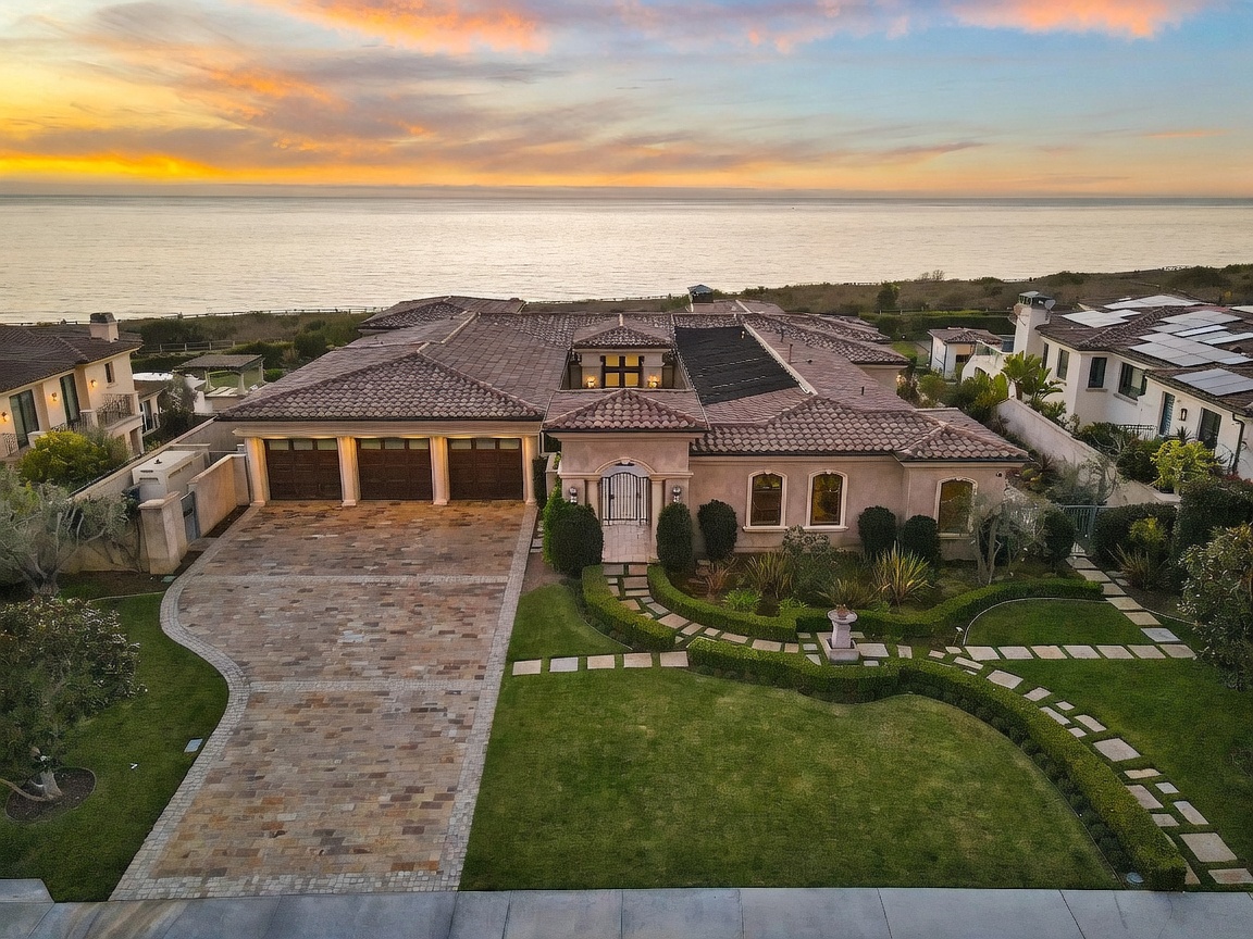 This high-angle aerial view captures a luxurious coastal residence at sunset, showcasing its expansive tile roof and multi-car garage. The property features a meticulously manicured lawn with stone pathways leading to the front entryway, all set against a stunning backdrop of the ocean horizon. The warm, golden light of the setting sun highlights the architectural details of the home and the surrounding landscape.
