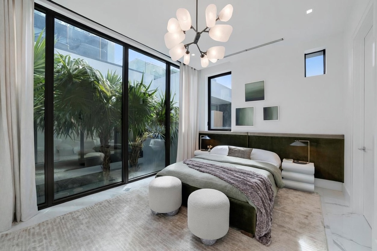 This modern bedroom features a plush bed with a deep green upholstered headboard and a neutral-toned textured rug. Large floor-to-ceiling windows provide an expansive view of a lush, private courtyard filled with tropical greenery. The room is accented by a unique, multi-bulb chandelier and minimalist bedside tables, creating a serene and sophisticated atmosphere.