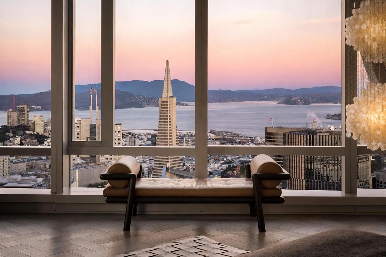 This interior space features a sophisticated, modern aesthetic centered around a luxurious leather daybed positioned before expansive floor-to-ceiling windows. The view showcases a breathtaking panoramic vista of the San Francisco skyline, including the iconic Transamerica Pyramid and the bay at sunset. A decorative, multi-tiered shell chandelier hangs to the right, adding a touch of elegance to the minimalist, high-rise setting.