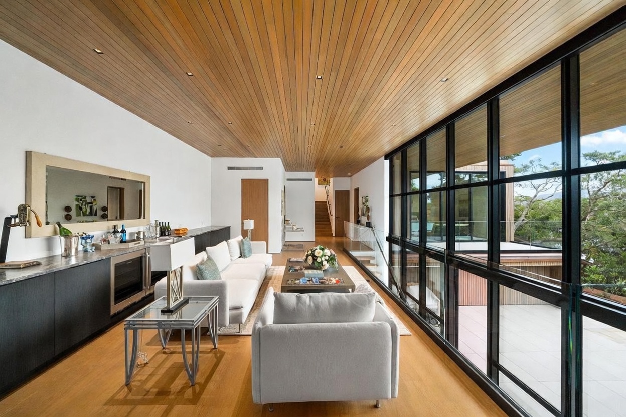 This sophisticated living room features a warm wood-paneled ceiling and light wood flooring, creating a cohesive and inviting atmosphere. A long, sleek bar area with dark cabinetry and a mirror backsplash runs along the left wall, while a wall of floor-to-ceiling windows on the right provides an expansive view of the surrounding trees and outdoor deck. The space is furnished with a plush white sofa and a matching armchair, centered around a low coffee table, emphasizing a clean, contemporary aesthetic.