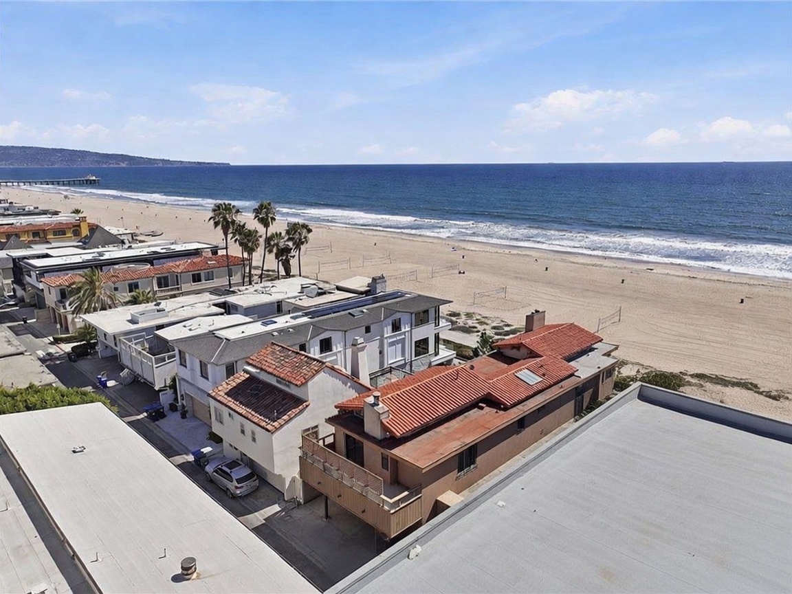 This high-angle aerial view captures a prime beachfront property situated directly along the sandy coastline. The scene highlights the proximity to the ocean, featuring a row of multi-level residential homes with varied rooflines and balconies overlooking the beach. The expansive blue horizon and the rhythmic waves meeting the shore create a serene and highly desirable coastal atmosphere.