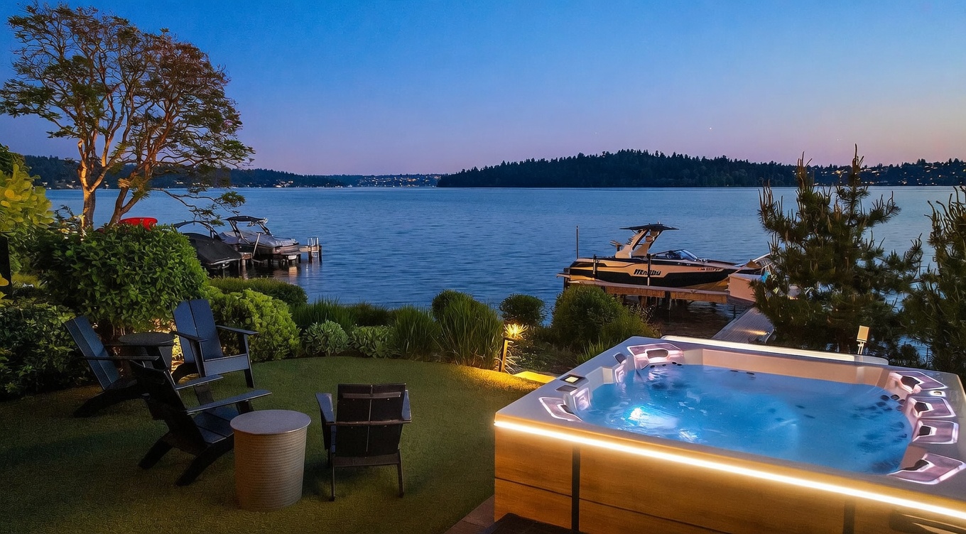 This serene waterfront scene features a modern, illuminated hot tub positioned on a manicured lawn overlooking a calm lake at dusk. The foreground includes a set of dark outdoor chairs and a small table, while the background showcases a private dock with two boats moored in the water. The twilight sky creates a peaceful, luxurious atmosphere, highlighting the property's prime location and outdoor living amenities.