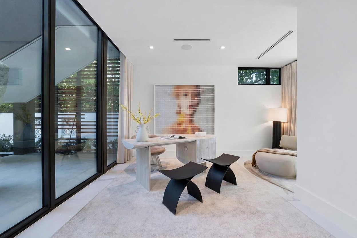 This modern, minimalist home office features a light-toned desk paired with two sculptural black stools, set against a large abstract art piece. The room is bathed in natural light from expansive floor-to-ceiling windows that look out onto a private outdoor area, while a curved lounge chair provides a cozy seating nook. The clean white walls and neutral area rug create a serene, sophisticated atmosphere suitable for a professional workspace.