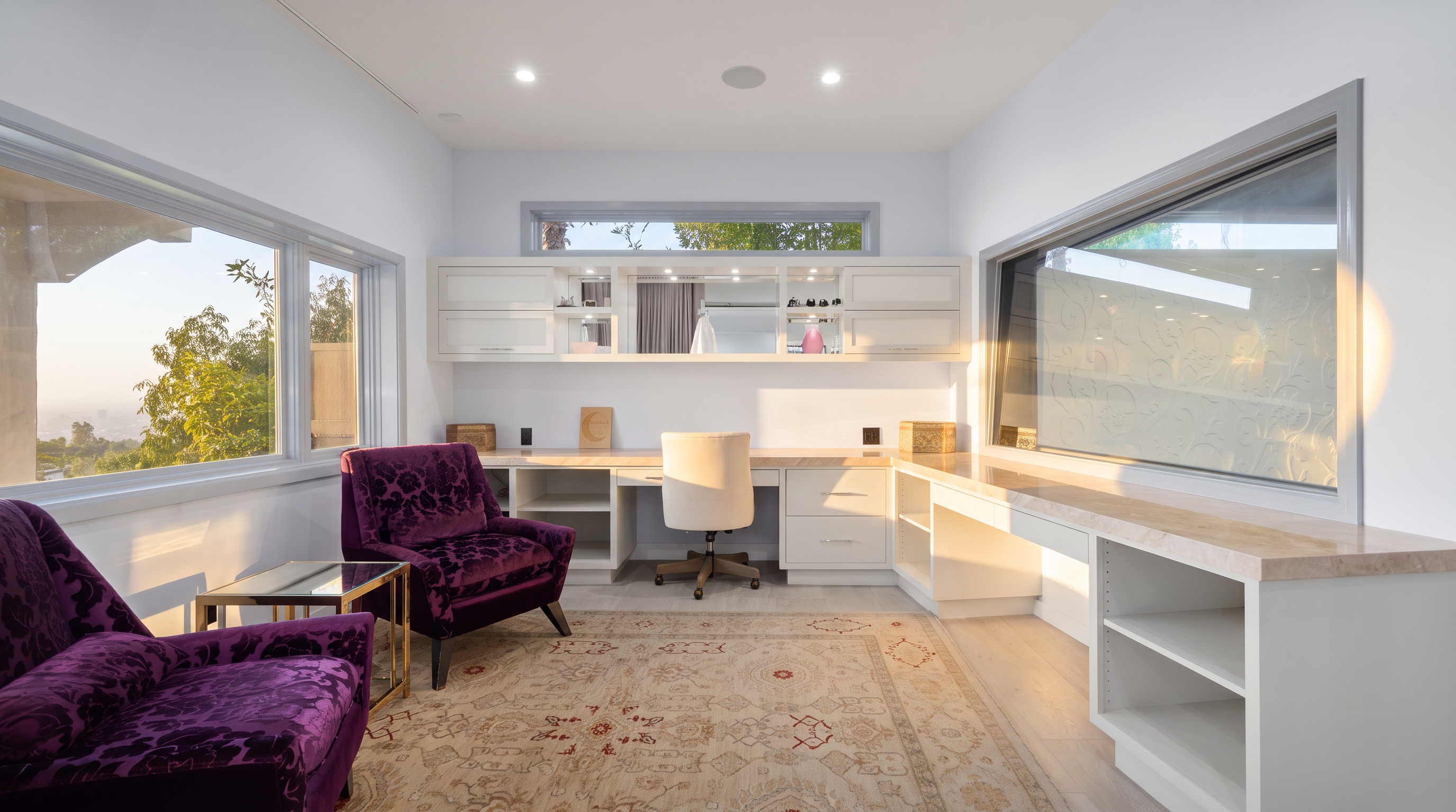 This bright and airy home office features a large, L-shaped built-in desk with a light-toned stone surface and ample white cabinetry for storage. Two vibrant purple velvet armchairs provide a comfortable seating area atop a large, intricately patterned area rug. The room is illuminated by recessed lighting and natural light from large windows on two sides, offering a serene and productive atmosphere.