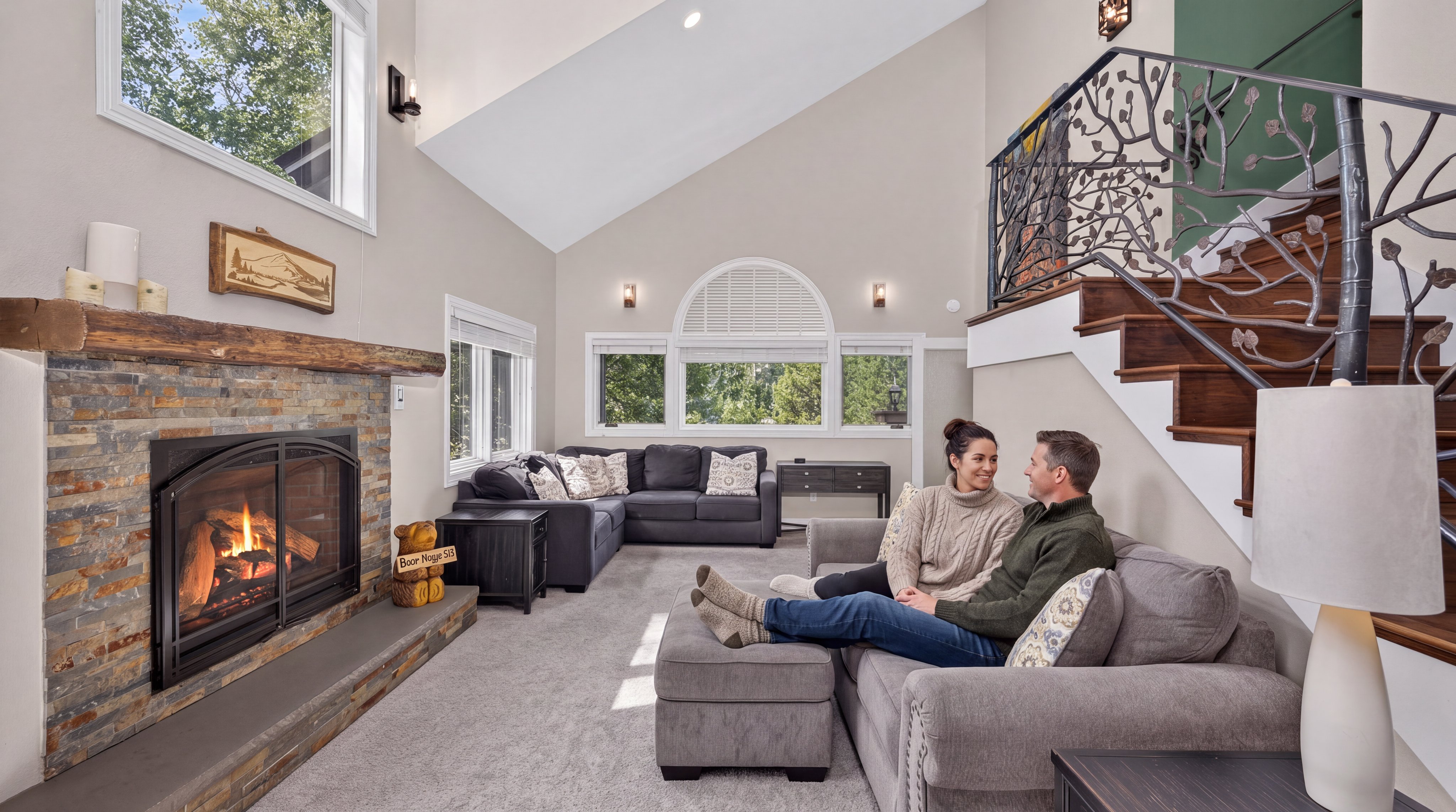 This cozy and inviting living room features a striking stone fireplace with a rustic wood mantel as its focal point, complemented by high vaulted ceilings and large windows that provide natural light. A comfortable sectional sofa and ottoman area, occupied by a couple, anchor the room, which also showcases an elegant staircase with a unique, artistic wrought-iron railing. The space blends rustic elements with modern comforts, creating a warm and well-appointed atmosphere ideal for relaxation.