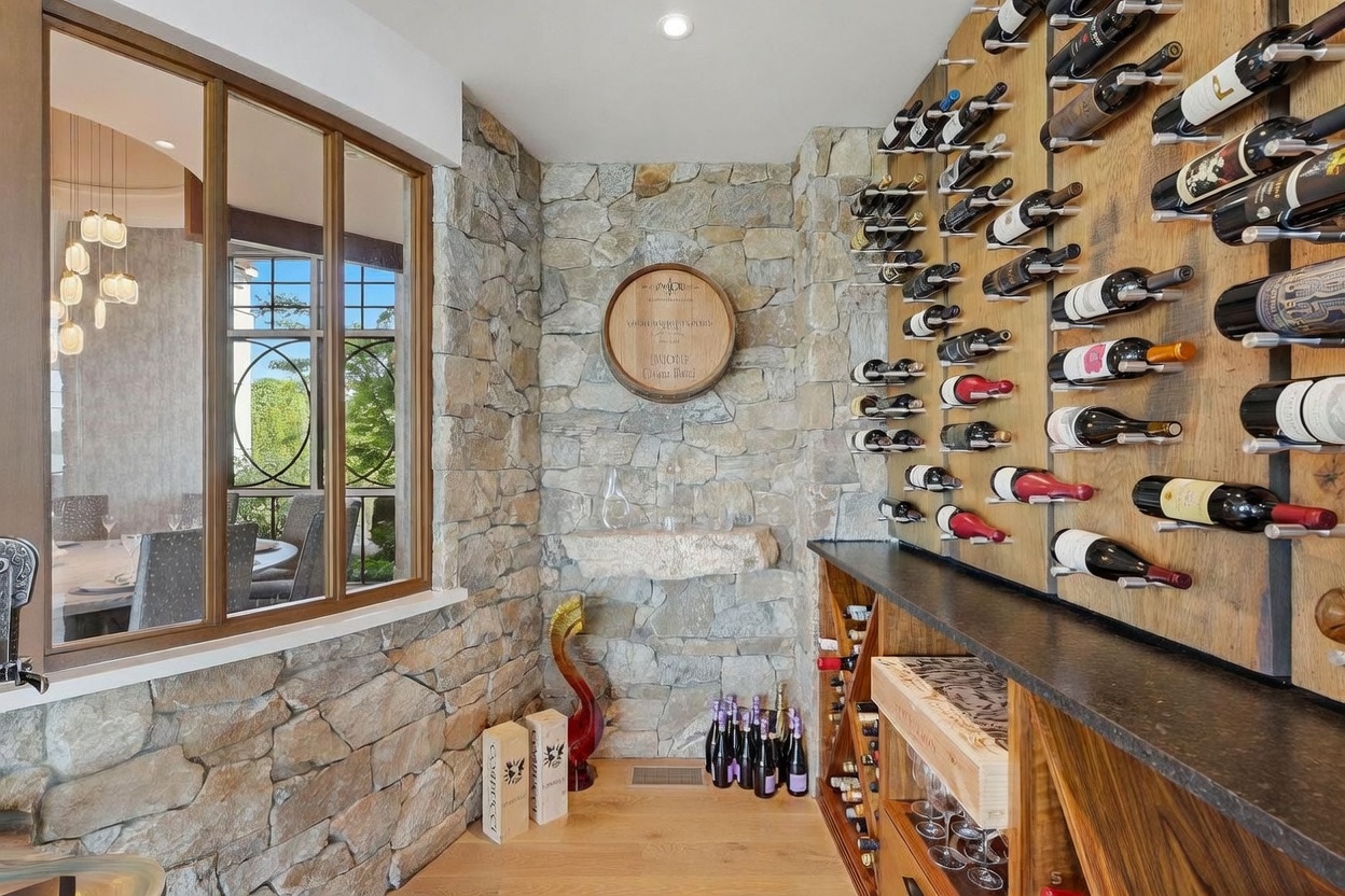 This sophisticated wine cellar features a rustic stone wall adorned with a decorative barrel head and a stone display shelf. A wall-mounted wine rack system showcases an extensive collection of bottles, while a glass partition provides a view into an adjacent dining area. The space is finished with light wood flooring and includes additional storage crates on the floor, creating an inviting and organized atmosphere for wine enthusiasts.