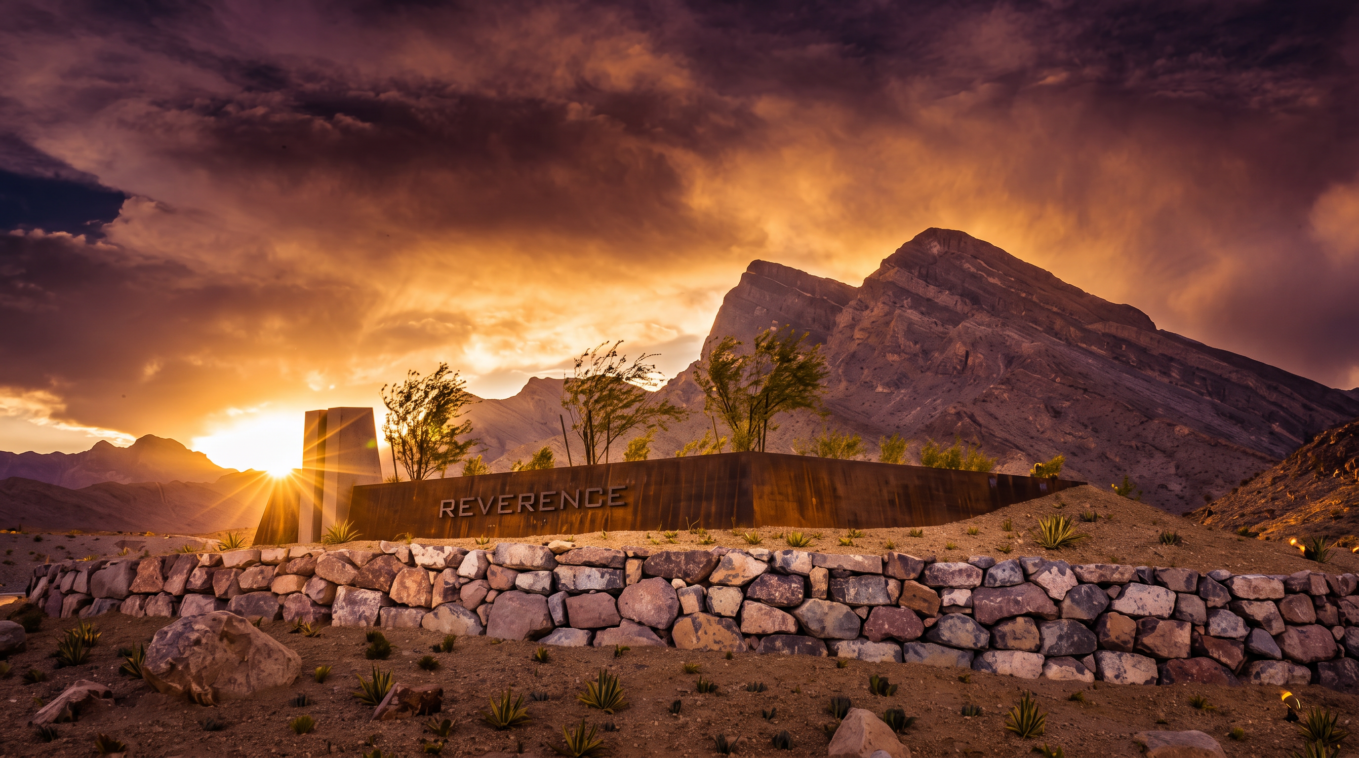 This image showcases the impressive entrance monument for the Reverence community, set against a dramatic, mountainous desert landscape at sunset. The structure features modern, weathered metal signage atop a textured stone retaining wall, highlighting the community's upscale and serene aesthetic. The scene effectively emphasizes the connection between the development and the natural beauty of its surroundings.