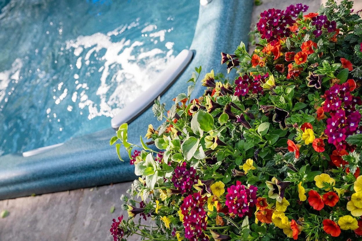 This close-up shot captures a vibrant, overflowing planter filled with a mix of purple, yellow, and orange flowers, positioned directly beside the edge of a bubbling hot tub. The textured, blue-toned spa shell contrasts with the lush, colorful foliage, creating a relaxing and inviting outdoor atmosphere. The perspective is intimate, focusing on the juxtaposition of natural beauty and leisure amenities.