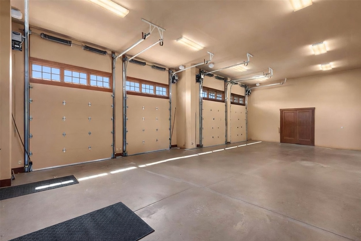This spacious, multi-bay garage features high ceilings and multiple overhead garage doors with integrated windows, allowing for ample natural light. The interior is finished with smooth, neutral-toned walls and a clean concrete floor, providing a versatile and well-maintained space. A set of double wooden doors is positioned on the far wall, adding a touch of warmth to the industrial aesthetic.