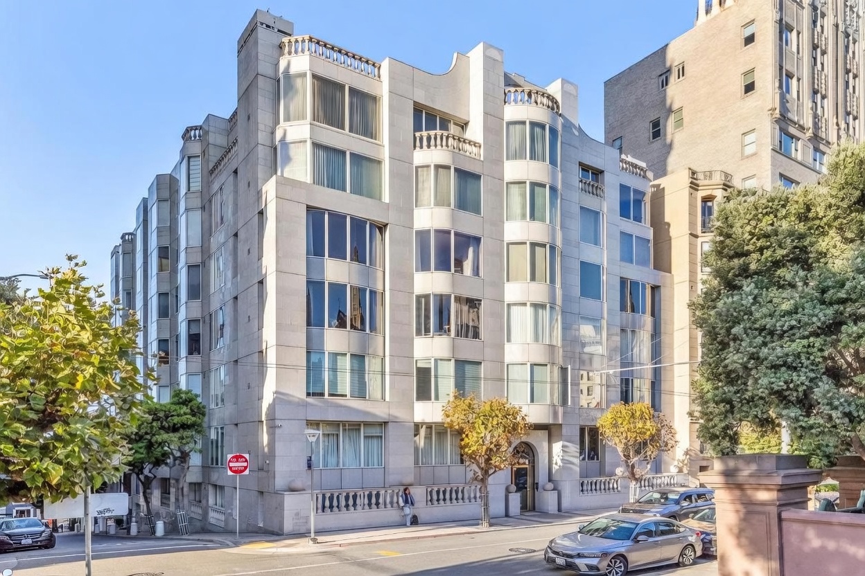 This image captures the front facade of a multi-story, light-colored stone apartment building in an urban setting. The architecture features a series of rounded, projecting window bays and a decorative balustrade along the roofline, conveying a sophisticated, classic aesthetic. A street-level view shows a sidewalk, a small tree, and parked cars, providing a sense of the building's integration into the neighborhood.