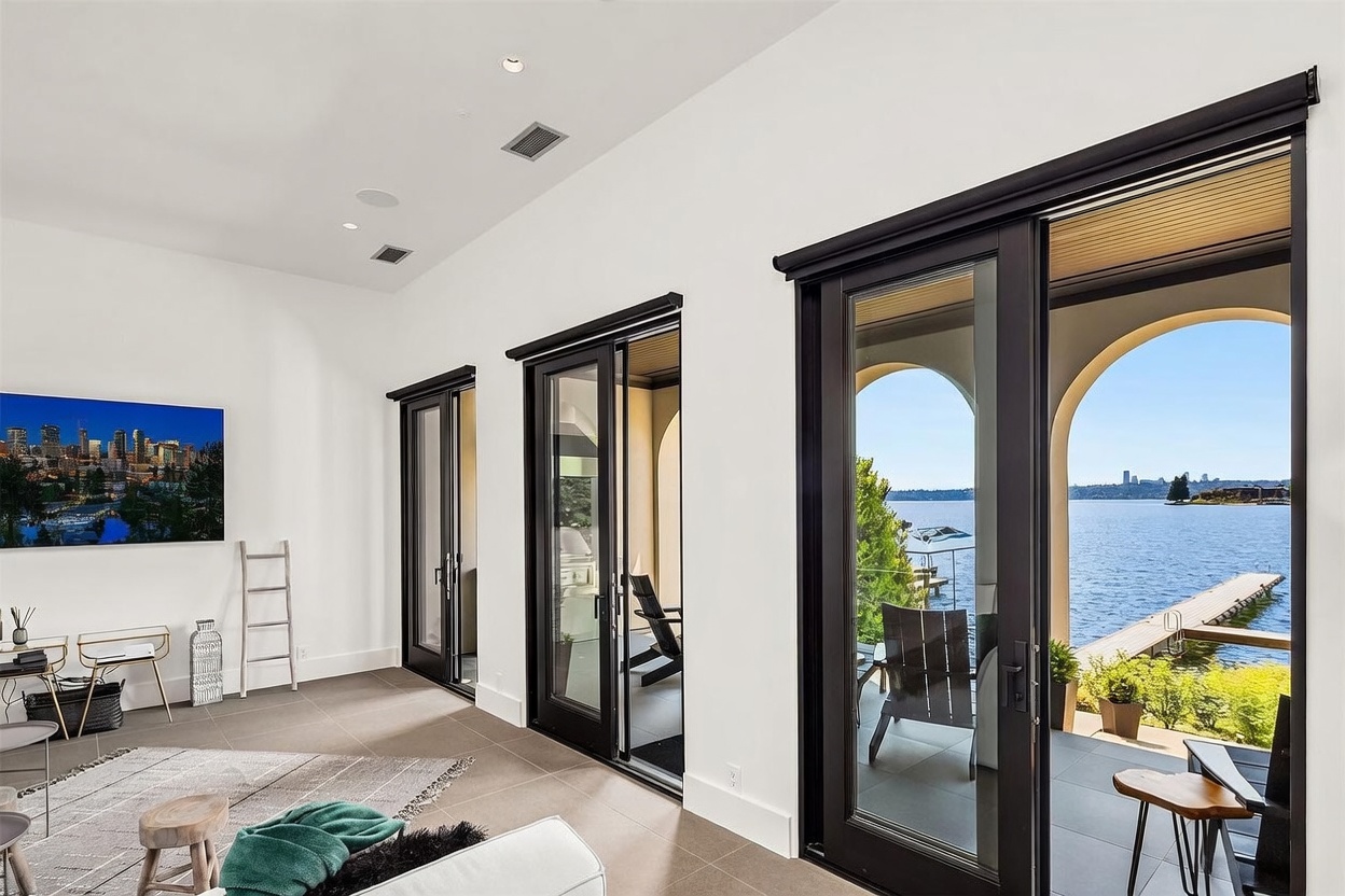 This bright, modern living space features large glass sliding doors that open onto a covered patio, framing a scenic view of the lake and distant city skyline. The room is finished with neutral-toned tile flooring, white walls, and recessed lighting, creating an airy and minimalist atmosphere. A television is mounted on the wall, and the space is furnished with a small wooden stool and a decorative ladder, emphasizing a clean, contemporary aesthetic.