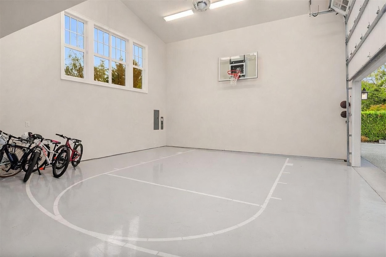 This spacious, high-ceilinged garage has been converted into a multi-purpose recreational area, featuring a basketball hoop mounted on the wall and painted court markings on the floor. The space is bright and clean, with large windows allowing for natural light and a row of bicycles parked neatly along the side wall. The perspective is from the open garage door, looking into the expansive, well-maintained interior.