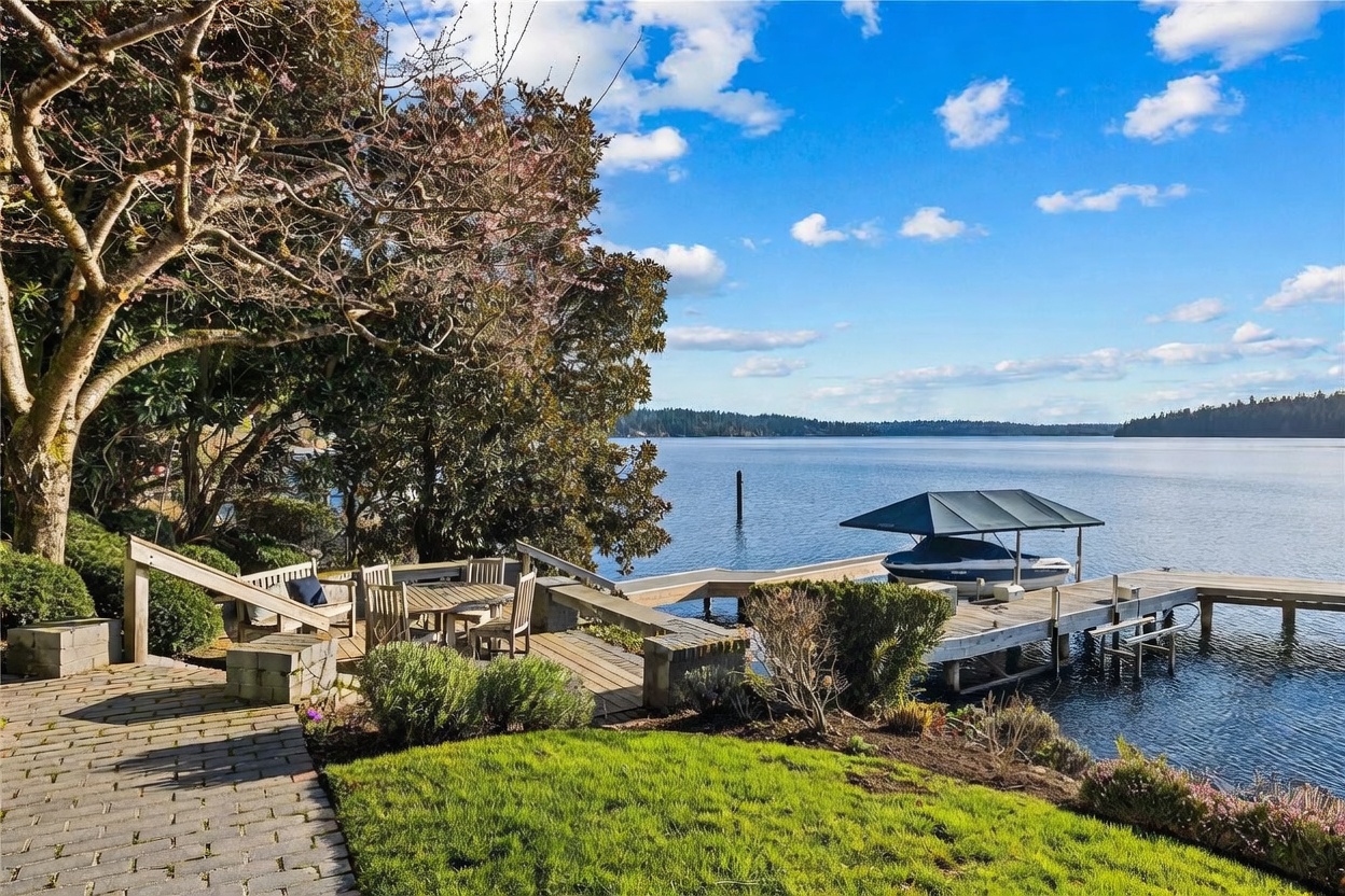 This scenic outdoor space features a multi-level wooden deck overlooking a calm lake, complete with a private dock and a covered boat slip. The area is surrounded by lush greenery and mature trees, creating a serene and private waterfront retreat. The perspective captures the inviting transition from the manicured lawn and stone pathway to the expansive water views.