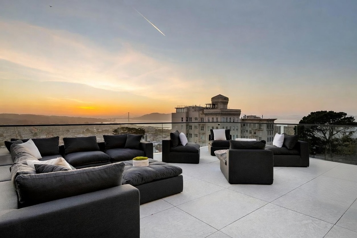 This expansive rooftop terrace offers a sophisticated outdoor living space with panoramic views of the city skyline and the Golden Gate Bridge at sunset. The area is furnished with modular dark-toned lounge seating arranged for conversation, set against a backdrop of glass railings that maintain an unobstructed view. The clean, modern aesthetic is complemented by light-colored stone pavers, creating a luxurious and serene atmosphere perfect for evening entertaining.
