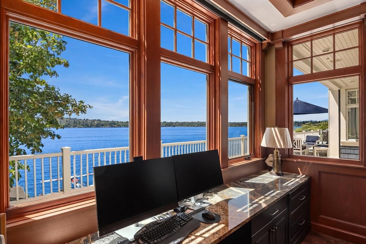This home office features a built-in granite desk positioned directly in front of expansive, floor-to-ceiling windows that offer a breathtaking, unobstructed view of a serene lake. The warm wood trim and cabinetry create a sophisticated, classic atmosphere, while the dual-monitor setup suggests a functional workspace. The perspective captures the seamless blend of professional productivity and tranquil natural beauty, highlighting the home's premium waterfront location.