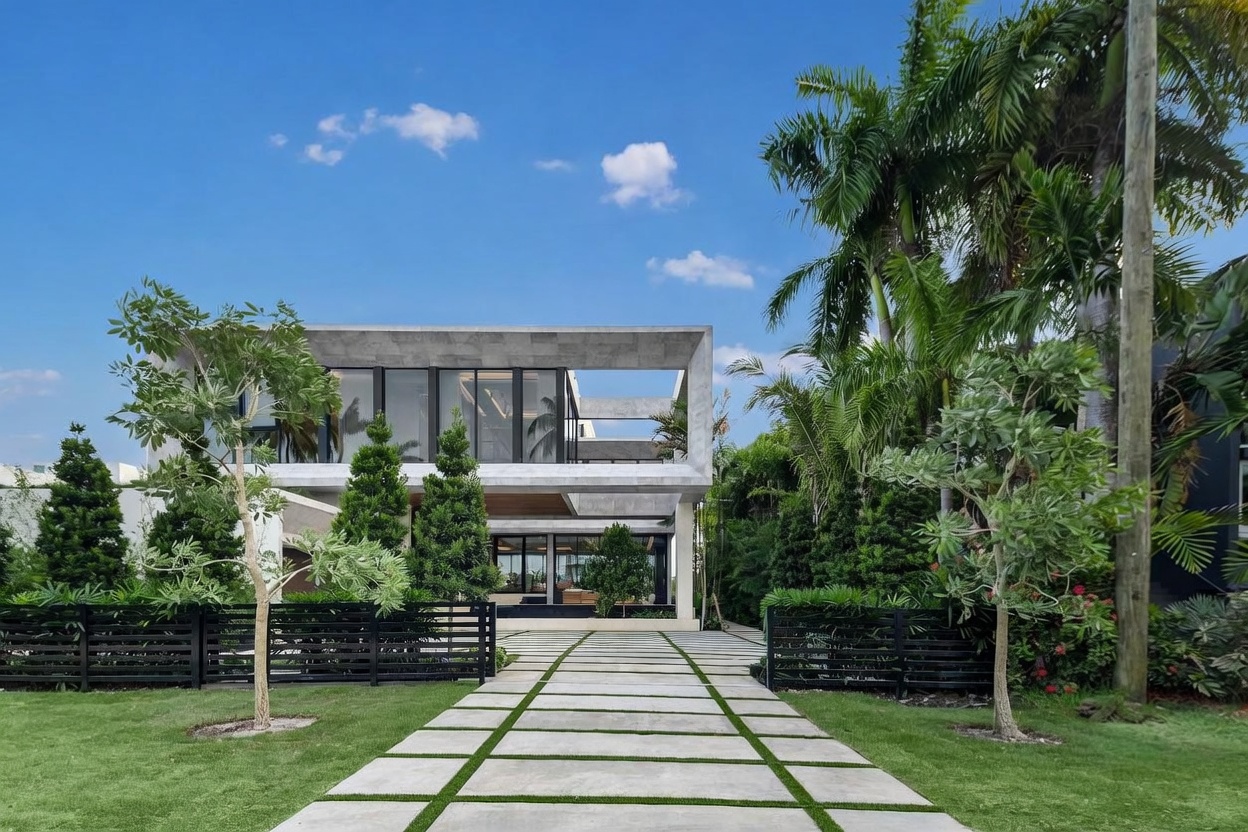 This striking modern residence features a bold, geometric architectural design with clean lines and a prominent concrete frame. The front approach is defined by a linear stone paver walkway flanked by manicured lawns, mature palm trees, and dark horizontal fencing. Large, expansive windows provide a clear view into the interior, emphasizing a seamless connection between the home's structure and the lush tropical landscaping.