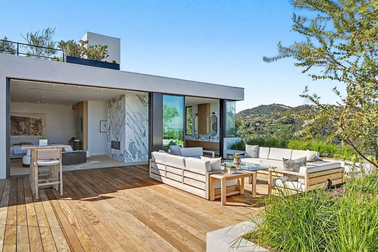 This expansive outdoor deck features light-toned wood planking and is furnished with comfortable, modern seating arrangements, including a sectional sofa and a high-top table. The space seamlessly transitions into an indoor living area through large sliding glass doors, revealing a striking stone-clad chimney breast. The setting offers a panoramic view of the surrounding hills, creating a serene and luxurious atmosphere for outdoor entertaining.