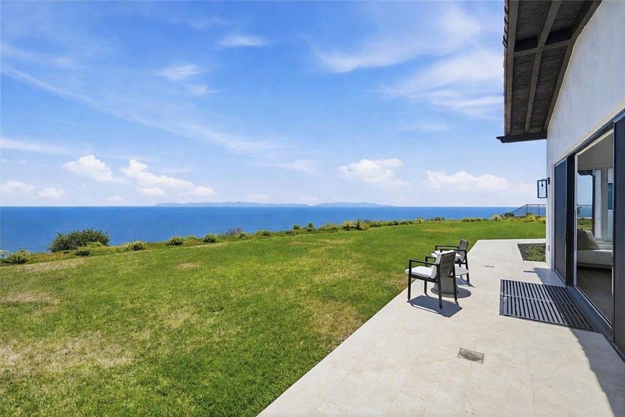 This image captures a spacious stone-paved patio overlooking a vast, manicured lawn that leads directly to a stunning ocean view. Two outdoor chairs are positioned on the patio, providing a serene spot to enjoy the expansive horizon and distant island silhouettes under a bright, clear sky. The perspective is from the edge of the home's exterior, highlighting the seamless transition between the indoor living space and the outdoor landscape.