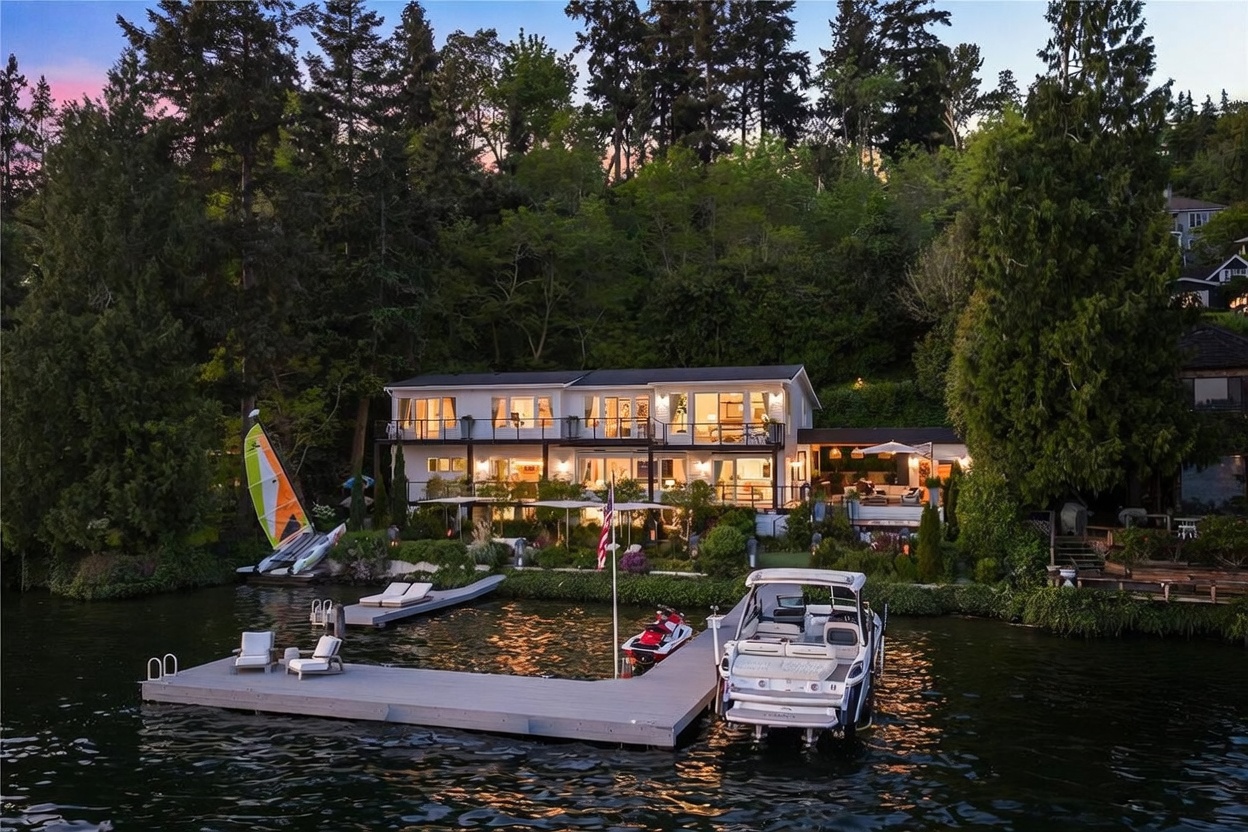This stunning waterfront property is captured from an elevated perspective, showcasing a multi-level home nestled against a lush, tree-covered hillside. The residence features expansive glass doors and balconies that overlook a private dock equipped with a boat and a jet ski, creating a serene and luxurious lakeside atmosphere. The warm glow from the interior lighting contrasts beautifully with the twilight sky, highlighting the home's seamless integration with its natural surroundings.