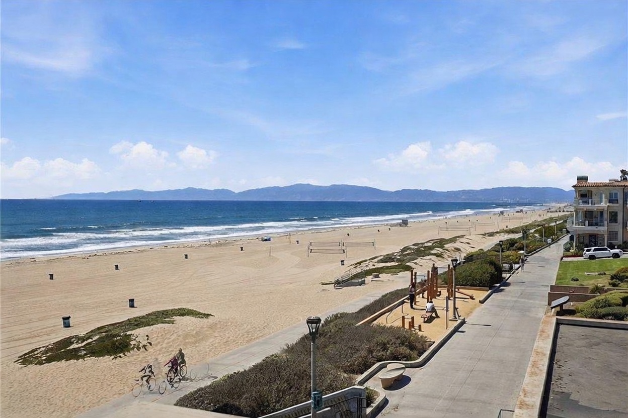This elevated perspective captures a sweeping, panoramic view of a sandy beach and the Pacific Ocean from a balcony. The scene includes a paved walkway, a small public playground area, and the vast coastline stretching toward distant mountains under a bright, clear sky. The vantage point emphasizes the property's prime location directly adjacent to the beach and recreational amenities.