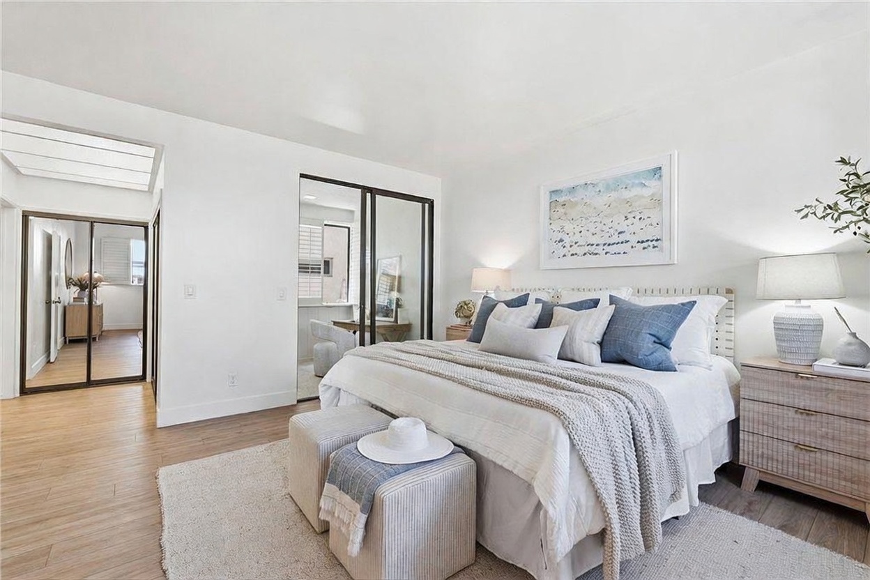This bright and airy primary bedroom features a neutral color palette with soft blue accents, centered around a plush bed dressed in layered linens and decorative pillows. The room includes a light wood nightstand, a pair of upholstered ottomans at the foot of the bed, and mirrored closet doors that reflect the adjacent space. A large piece of coastal-inspired artwork hangs above the headboard, contributing to the serene and inviting atmosphere of the room.
