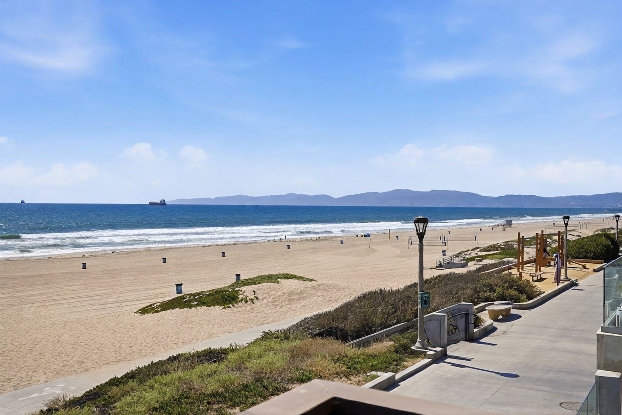This elevated balcony view captures a stunning, unobstructed panorama of the Pacific Ocean and a wide sandy beach. The perspective looks out over a paved walkway, a small patch of coastal vegetation, and a public beach area equipped with volleyball nets and playground structures. The bright, clear sky and expansive horizon create a serene and highly desirable coastal atmosphere.