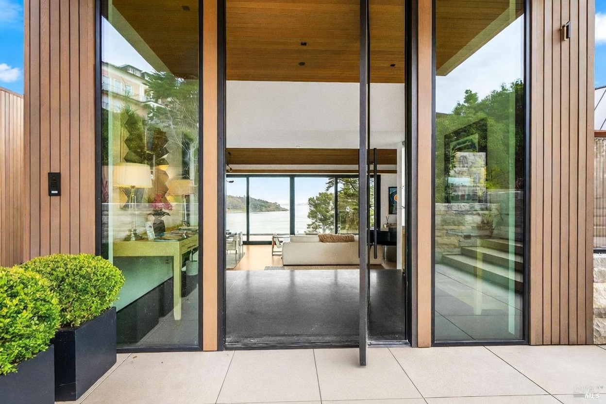 This modern entryway features a large, pivoting glass door framed by vertical wood siding and expansive glass panels. The perspective looks through the open doorway into a bright living space that offers a clear view of the water and distant hills. A textured doormat sits on the light-colored patio, while a potted shrub adds a touch of greenery to the left of the entrance.