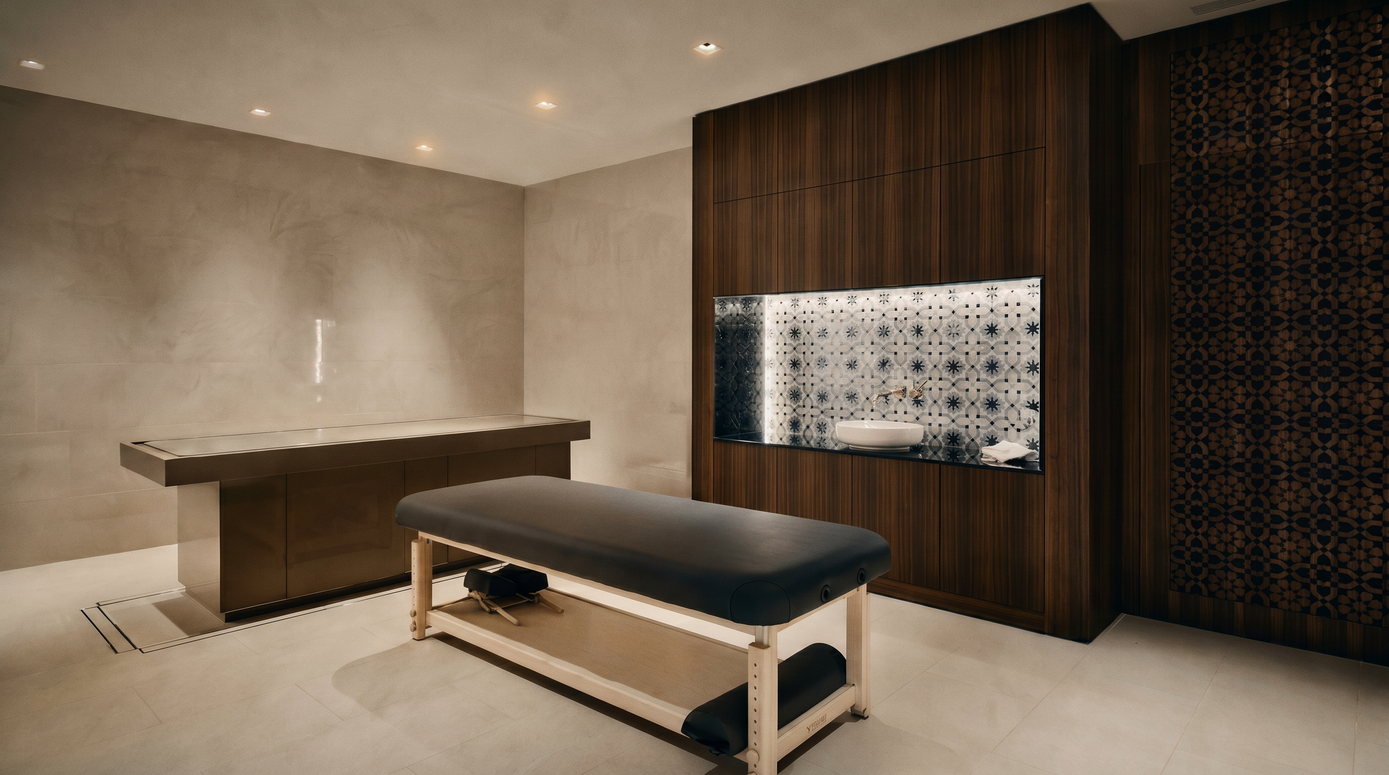 This image showcases a luxurious, spa-inspired massage room featuring a professional treatment table and a built-in vanity area with a decorative tiled backsplash. The room is characterized by warm wood paneling, recessed lighting, and a serene, minimalist aesthetic designed for relaxation. The perspective is a wide, eye-level shot that captures the functional layout and high-end finishes of this private wellness space.