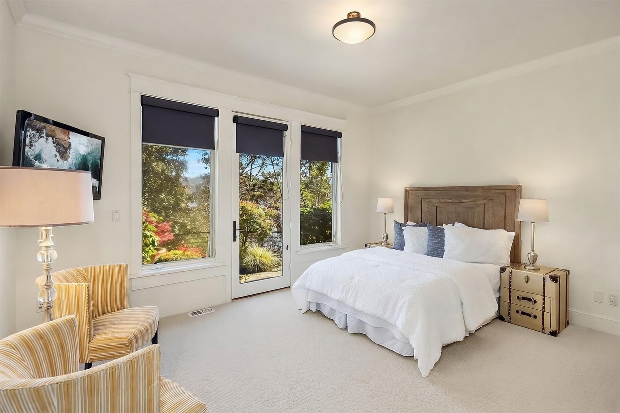 This bright and airy primary bedroom features a plush white bed with a wooden headboard, flanked by two unique, trunk-style nightstands. A cozy seating area with two striped armchairs sits near a large window and glass door that provide abundant natural light and a view of the lush outdoor greenery. The room is finished with neutral carpeting and a simple, elegant ceiling light fixture, creating a serene and inviting atmosphere.