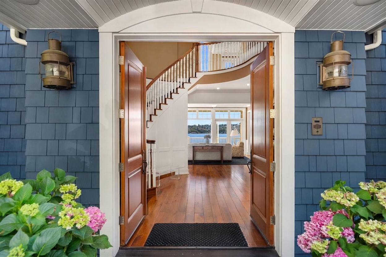 This inviting entryway features double wooden doors standing open to reveal a bright, elegant interior with a grand staircase and a stunning view of the water beyond. The exterior is clad in classic blue shingles, accented by traditional nautical-style lanterns on either side of the doorway. The perspective is framed from the threshold, drawing the viewer's eye through the home toward the scenic outdoor vista.