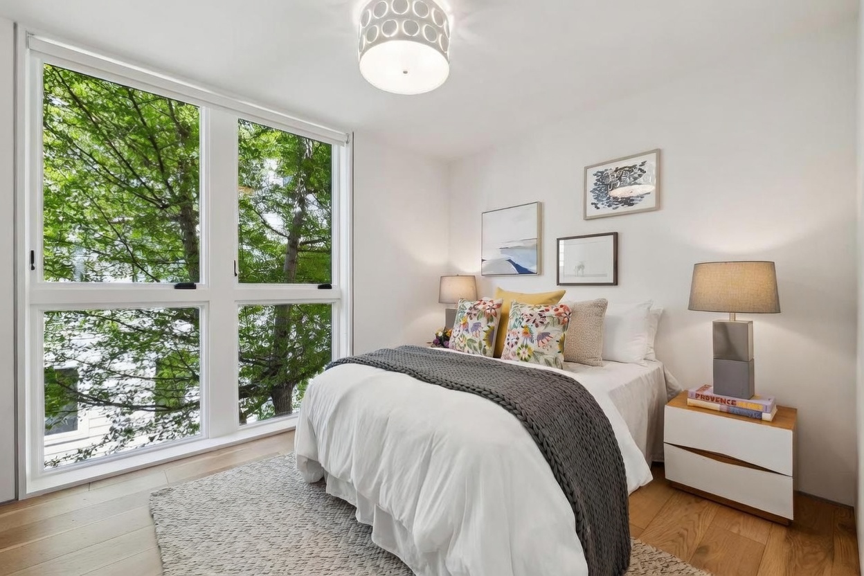 This bright and airy bedroom features a queen-sized bed dressed in white linens with a textured gray throw and colorful accent pillows. Large windows on the left wall provide an expansive view of lush green trees, filling the space with natural light. The room is accented by a modern drum pendant light, a light wood nightstand with a contemporary lamp, and a collection of framed art on the wall.