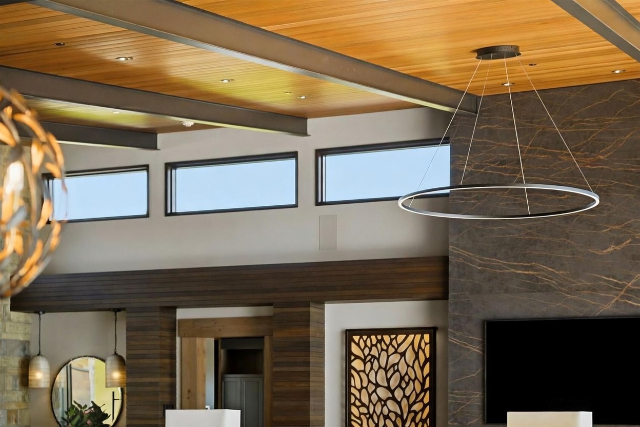 This interior view showcases a sophisticated living space with a warm wood-paneled ceiling accented by exposed structural beams. A modern, circular chandelier hangs prominently in front of a dark, textured stone wall, while a series of horizontal windows allows natural light to filter into the room. The design aesthetic is contemporary and refined, featuring clean lines and a mix of rich wood tones and dark, dramatic surfaces.
