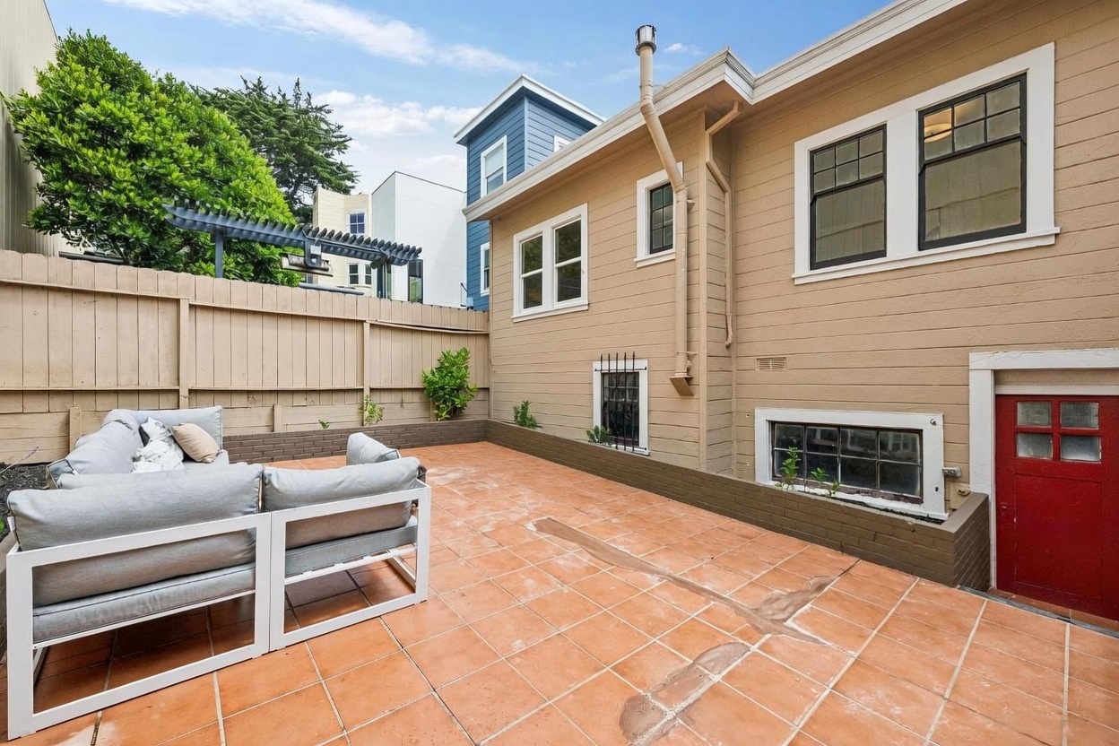 This image captures a private, paved patio area adjacent to the rear of a multi-story home. The space features a modern, light-gray outdoor sectional sofa set and is enclosed by a tall wooden privacy fence, creating a secluded atmosphere. The exterior of the house is finished in tan siding with white-trimmed windows and a distinct red door, providing a clean and well-maintained appearance.