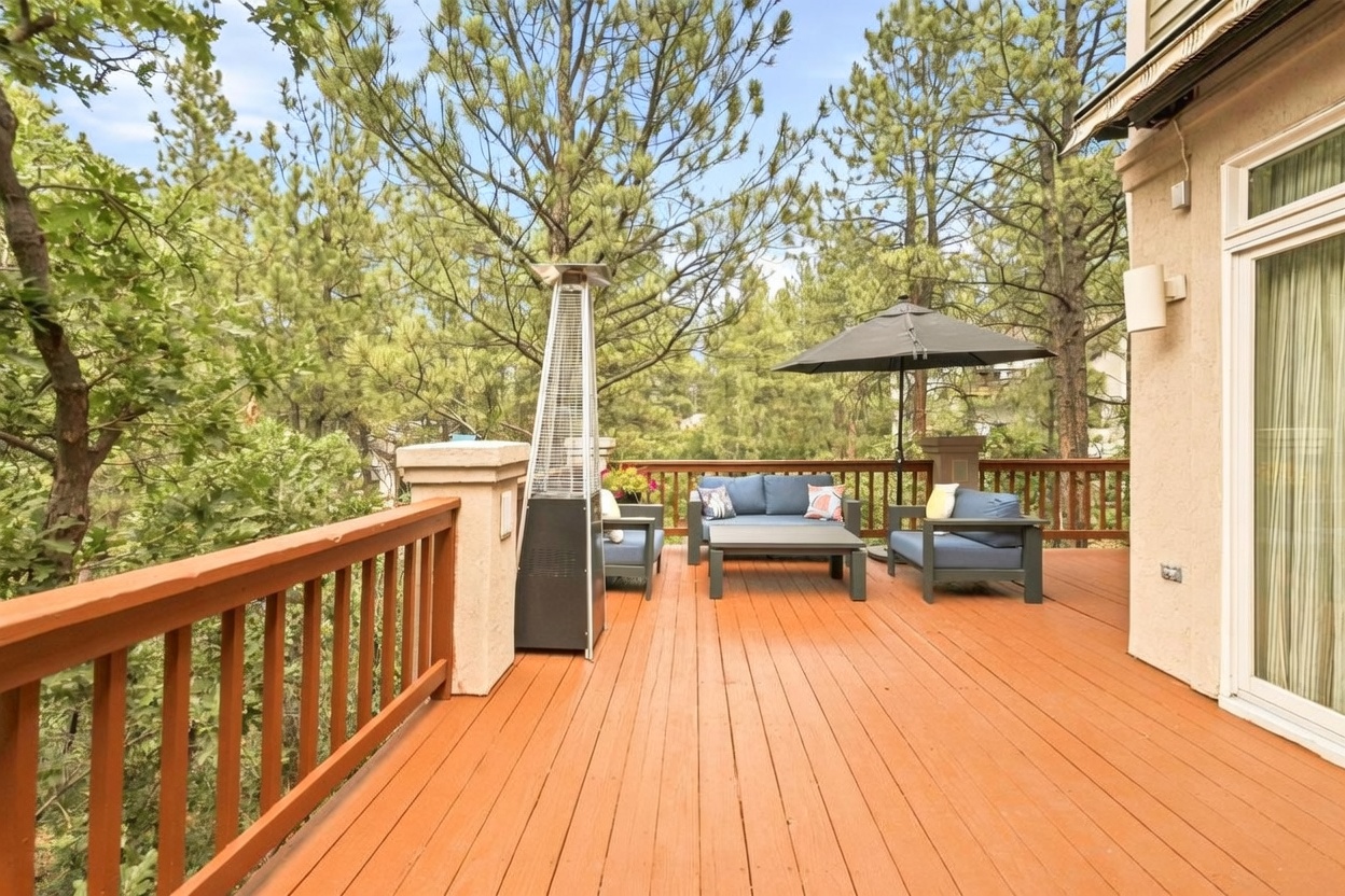 This elevated wooden deck offers a serene outdoor living space surrounded by a lush forest of pine trees. The area is furnished with a comfortable blue seating set, a central coffee table, and a tall outdoor heater, all shaded by a dark patio umbrella. The perspective captures the inviting, private atmosphere of the deck, which connects directly to the home's interior through large glass sliding doors.