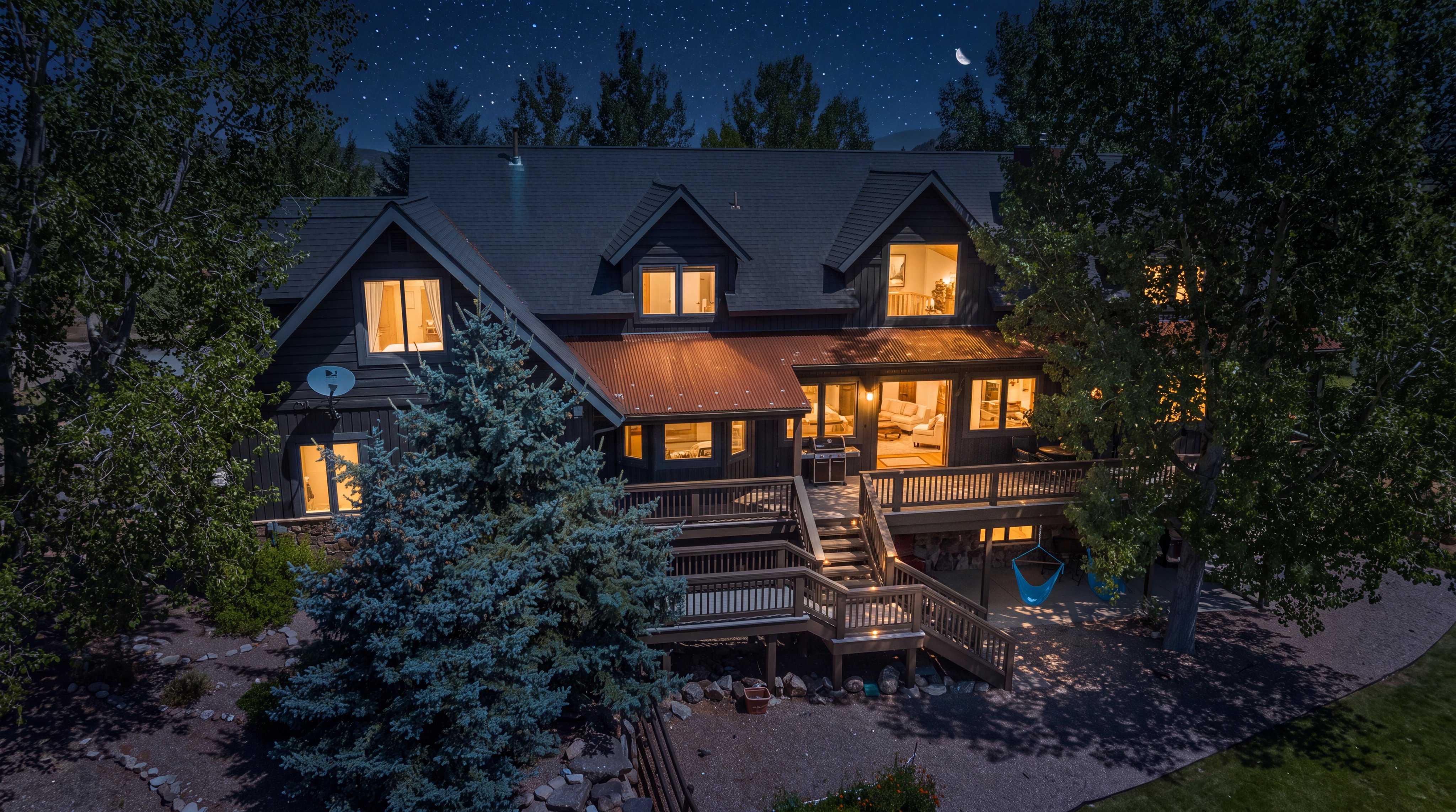 This is an elevated aerial perspective of a sprawling rustic-style home with dark wood siding, a distinctive copper-colored metal roof section, and multiple gables. The property features an expansive multi-level wooden deck with staircase access and is surrounded by mature trees and natural, landscaped grounds. The combination of rural charm and functional outdoor living space creates a welcoming and picturesque mountain home aesthetic.