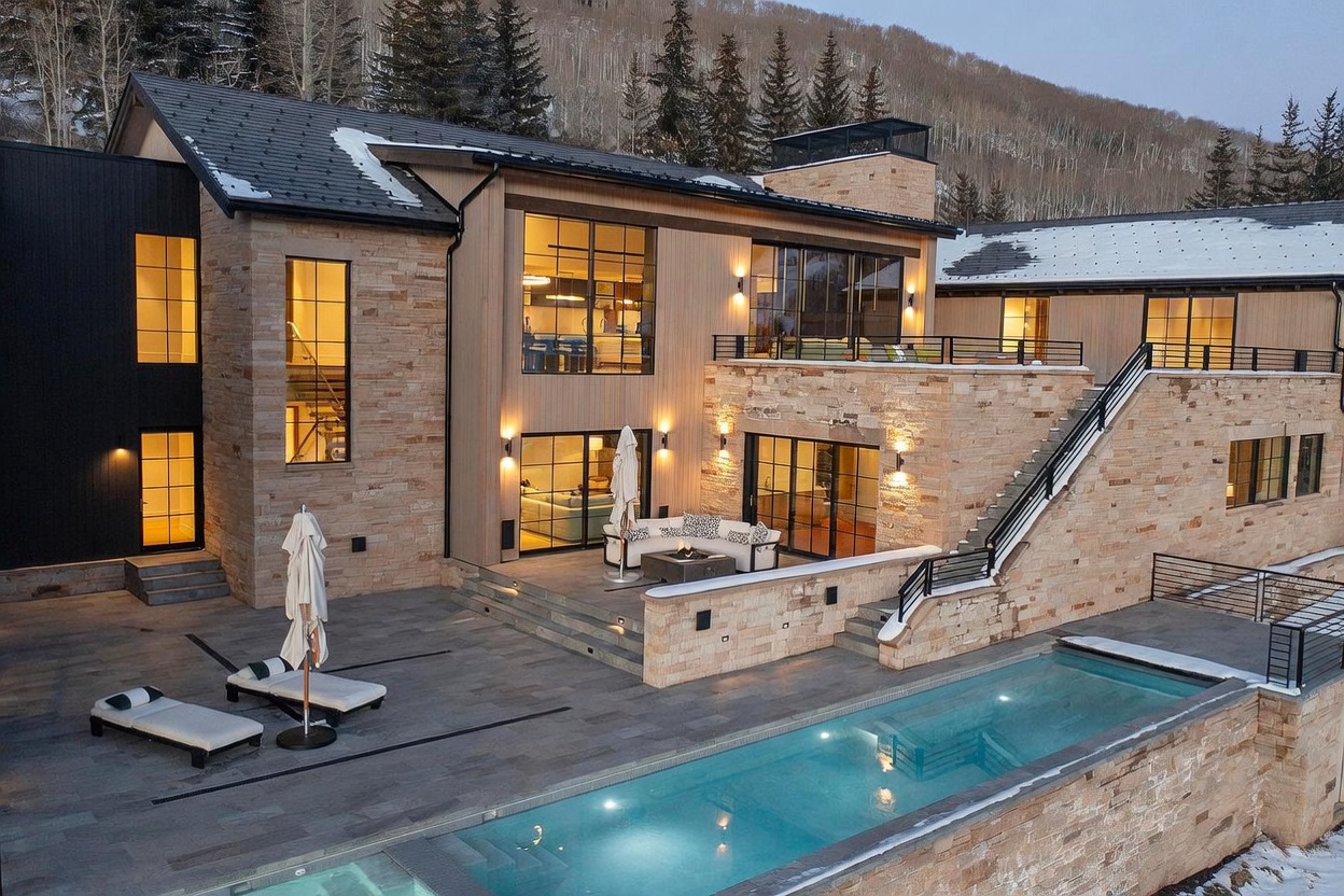 This luxurious mountain estate features a multi-level exterior design with a blend of natural stone and wood siding. The rear patio includes a comfortable seating area with a fire pit, overlooking a sleek, rectangular swimming pool that stretches across the terrace. Large windows and glass doors provide a seamless connection between the indoor living spaces and the expansive outdoor entertainment area, all set against a backdrop of snow-dusted pine trees.