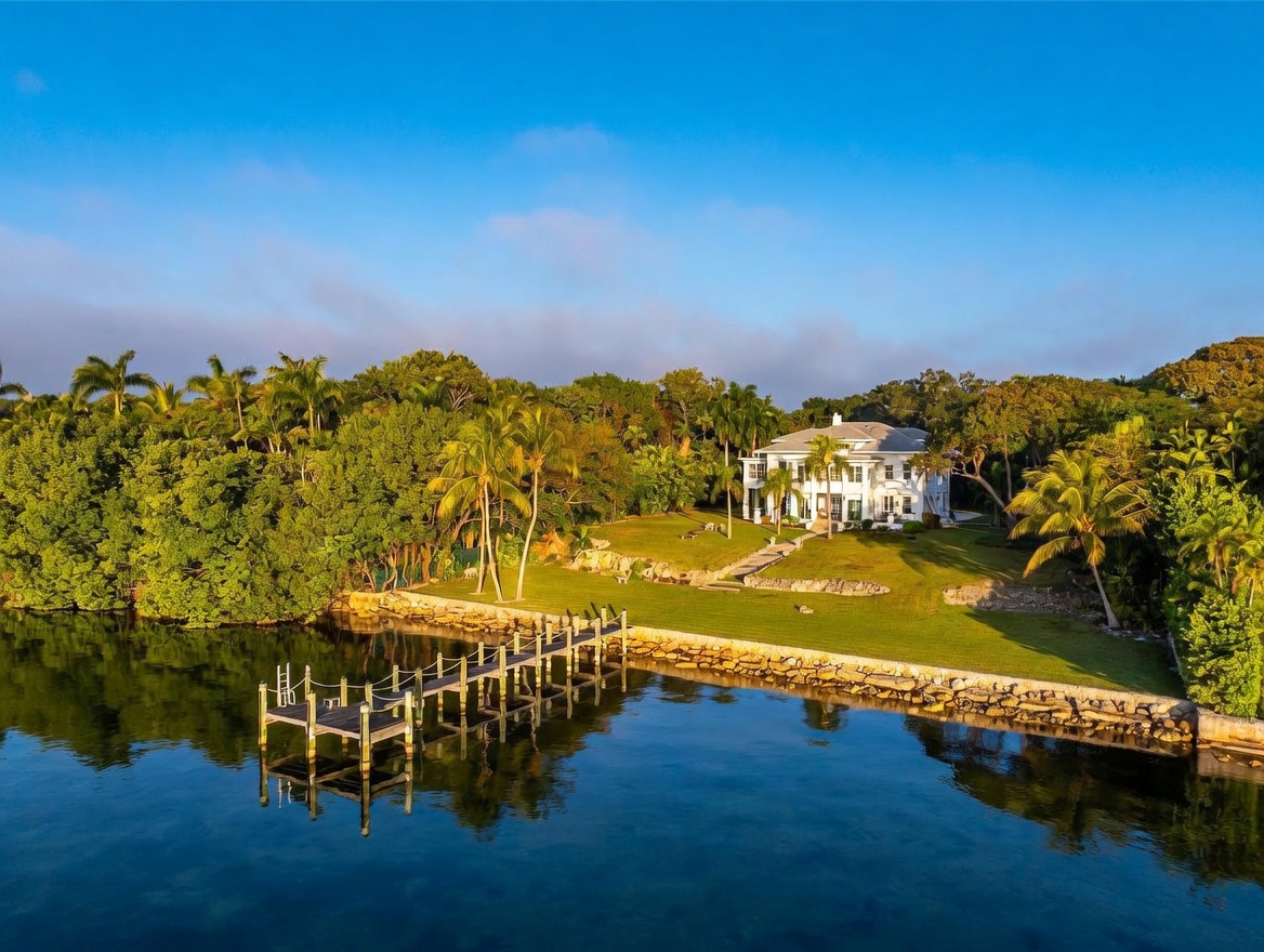 This aerial perspective captures a grand, multi-story estate situated on a lush, expansive waterfront property. The home features a classic architectural style with a prominent white facade, surrounded by mature trees and a manicured lawn that slopes gently toward a private wooden dock extending into the calm water. The scene conveys a sense of serene luxury and privacy, highlighting the seamless integration between the residence and its natural coastal environment.