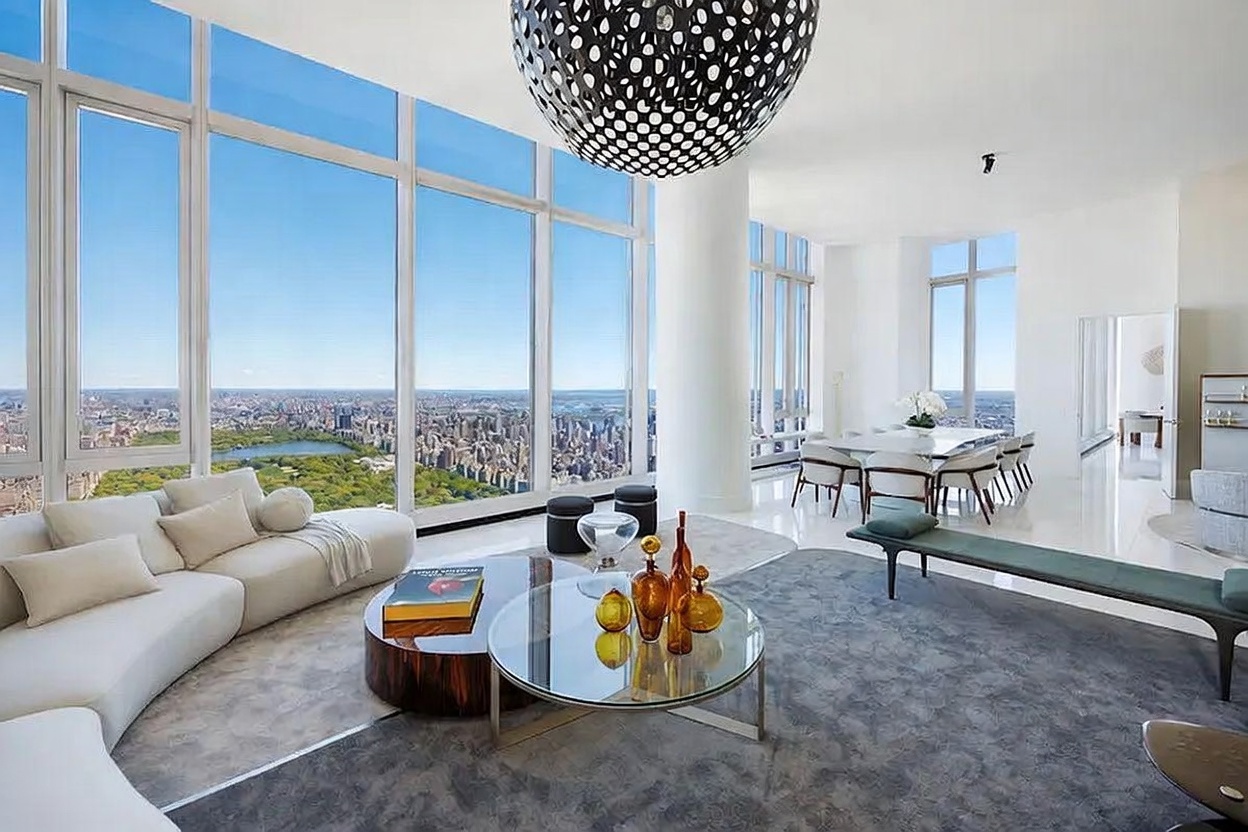 This expansive, high-rise living room features floor-to-ceiling windows that provide a panoramic view of the city skyline and a large park. The space is furnished with a curved, light-colored sofa, a circular glass coffee table, and a dark grey area rug, all centered beneath a striking, large black spherical pendant light. A dining area is visible in the background, maintaining the open-concept layout and bright, minimalist aesthetic.