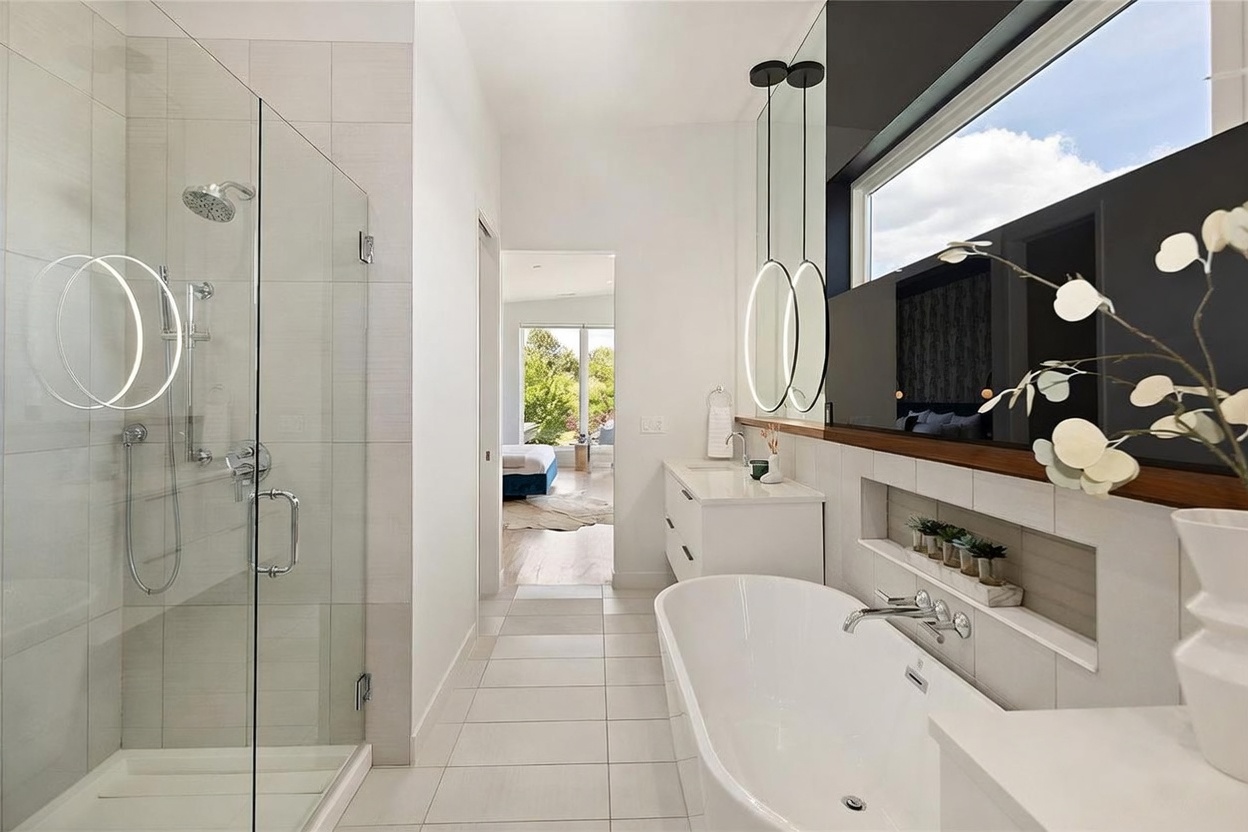 This modern primary bathroom features a sleek, glass-enclosed walk-in shower on the left and a freestanding soaking tub on the right. A double vanity with circular illuminated mirrors sits beneath a horizontal window, while a recessed niche provides convenient storage above the tub. The space is finished with light-toned tile flooring and walls, creating a bright, airy, and contemporary atmosphere that leads directly into an adjacent bedroom.