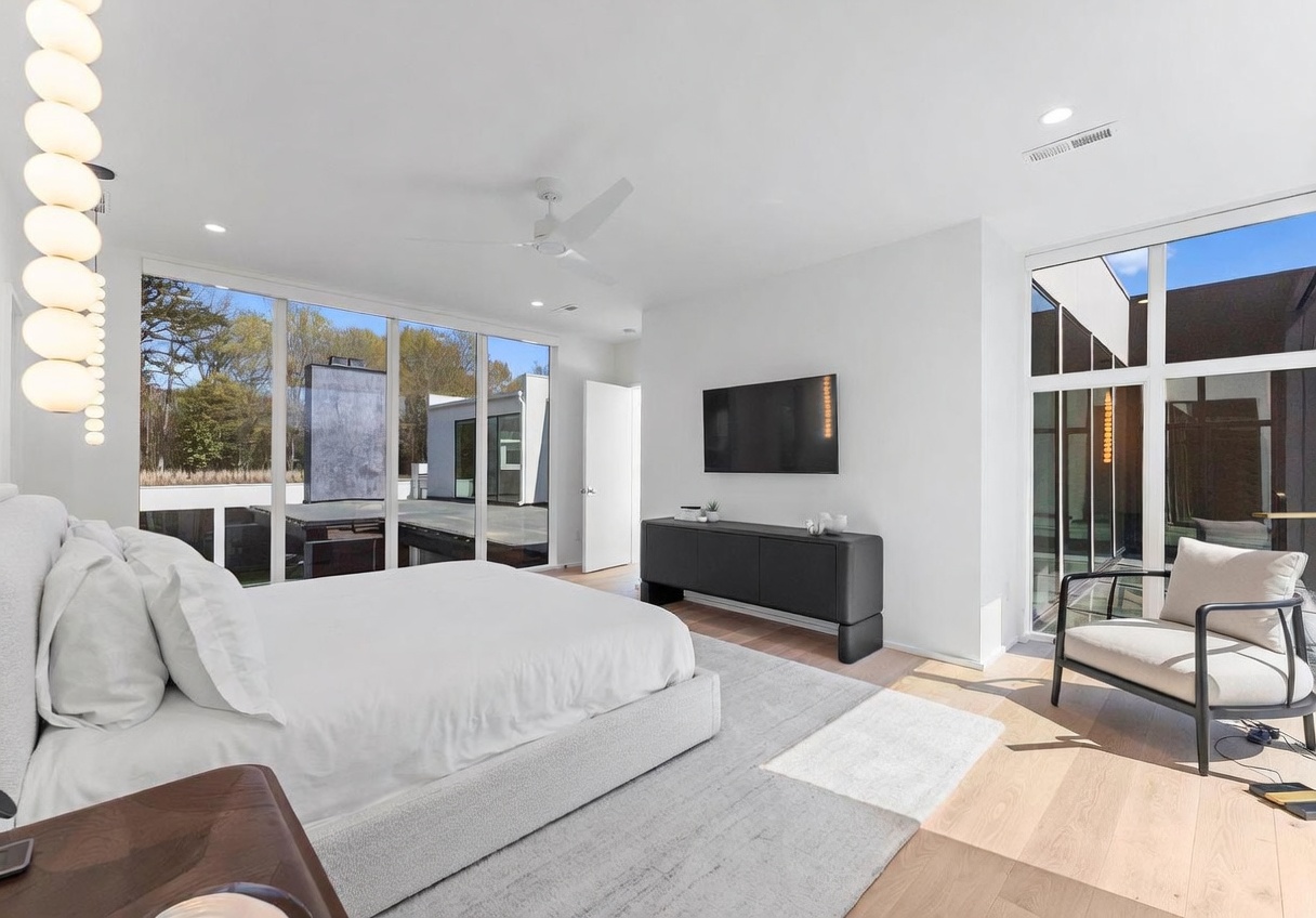 This modern primary bedroom features a minimalist aesthetic with a large, light-colored bed as the centerpiece, set against a backdrop of expansive glass walls that provide a seamless view of the outdoor deck. A sleek, dark console table sits beneath a wall-mounted television, while a contemporary armchair is positioned near the corner to create a cozy reading nook. The room is bathed in natural light, emphasizing the clean lines and neutral color palette of the space.