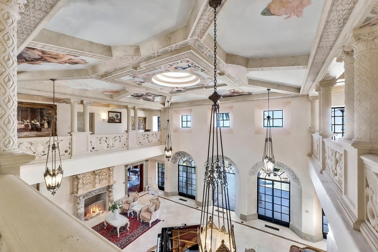 This grand, double-height living space features an ornate, painted ceiling with a central skylight and classical architectural detailing. The room is viewed from an elevated mezzanine, showcasing a stone fireplace with a decorative mantel, large arched windows, and multiple hanging chandeliers. The open-concept layout includes a seating area with a patterned rug and a grand piano, creating an atmosphere of opulence and refined elegance.