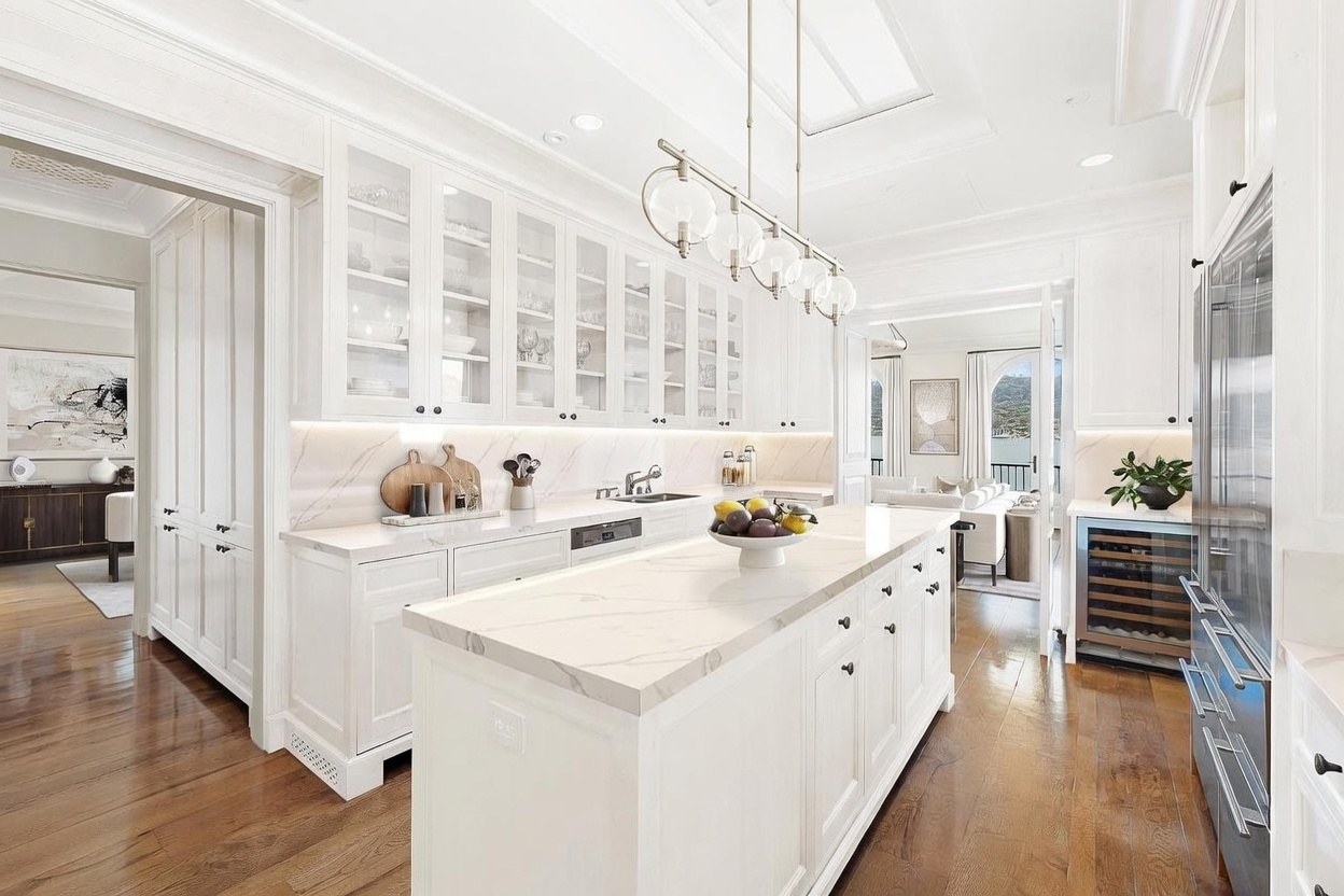 This bright, modern kitchen features crisp white cabinetry with glass-front upper cabinets and a large central island topped with light-veined stone. A multi-globe pendant light fixture hangs above the island, while a wine refrigerator is integrated into the cabinetry near the stainless steel appliances. The space flows seamlessly into an adjacent living area, creating an open and airy atmosphere with warm wood flooring throughout.