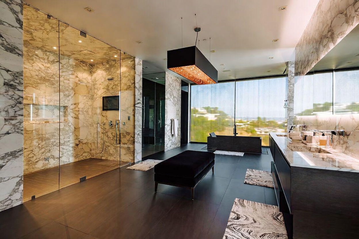 This luxurious primary bathroom features a sophisticated, modern aesthetic with floor-to-ceiling marble walls and dark wood flooring. The space is anchored by a sleek, dark freestanding bathtub positioned in front of a large window with a scenic view, complemented by a spacious glass-enclosed walk-in shower and a contemporary vanity. A central black velvet ottoman and a dramatic, textured pendant light fixture add a touch of opulence to the expansive, high-end design.