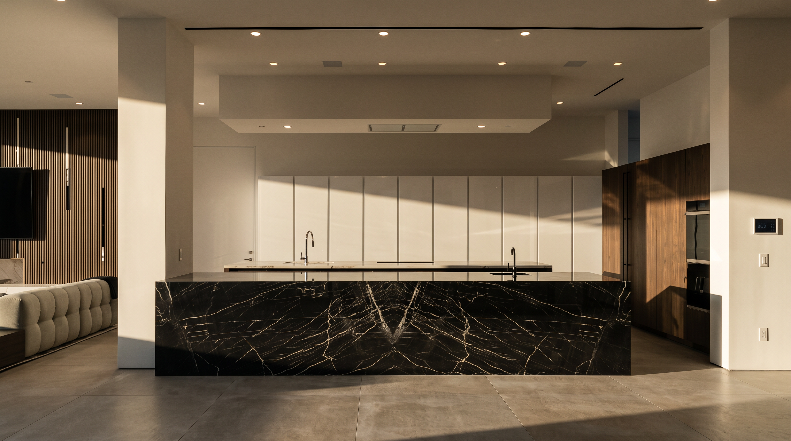 This expansive, modern kitchen features a prominent central island with a striking dark, veined stone finish. Behind the island, a long, minimalist counter with integrated sinks sits against a wall of sleek, white cabinetry that provides a clean, high-contrast aesthetic. The room is illuminated by recessed lighting and a large, suspended architectural soffit, creating a sophisticated and open atmosphere.