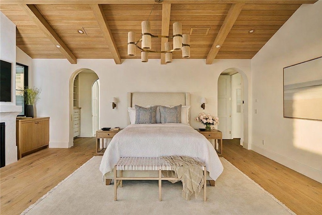 This primary bedroom features a striking vaulted ceiling with exposed light wood beams and a central cylindrical chandelier. The room is anchored by a neutral-toned bed with patterned accent pillows, flanked by matching light wood nightstands and arched doorways leading to adjacent spaces. A large area rug sits atop light wood flooring, creating a serene and sophisticated atmosphere.