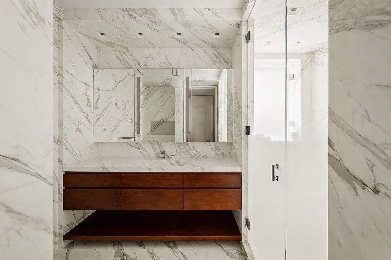 This modern bathroom features a floating vanity with dark wood cabinetry and a stone countertop, set against walls clad in light-toned, veined stone. A large, multi-panel mirror sits above the vanity, while a glass-enclosed shower stall is positioned to the right. The space is illuminated by recessed lighting, creating a clean, sophisticated, and high-end aesthetic.