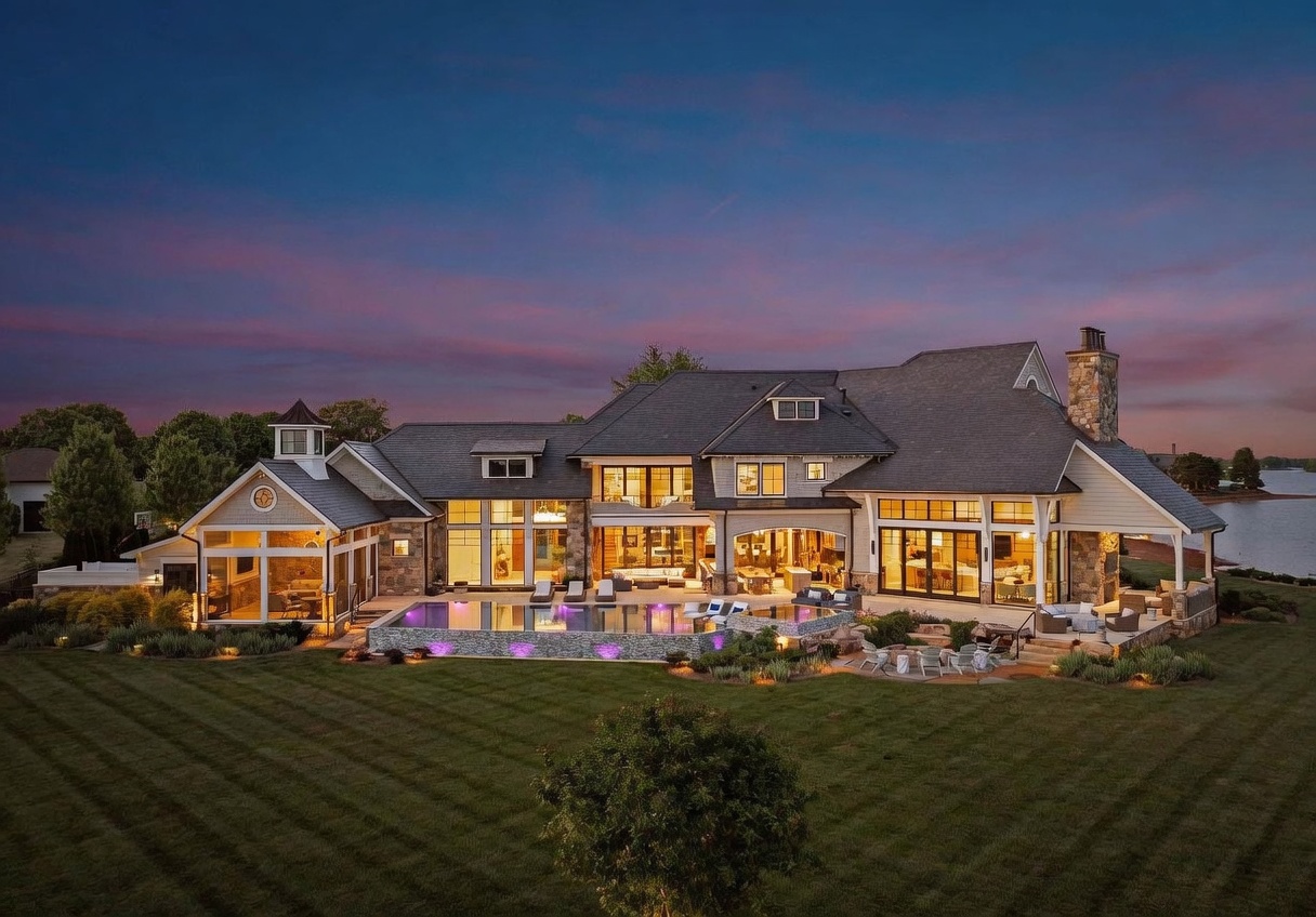 This expansive luxury estate is captured at twilight, showcasing a multi-level rear facade with a blend of stone and siding architecture. The property features a large infinity-edge pool with integrated lighting, a spacious stone patio, and a manicured lawn that leads toward a serene lakefront setting. The home's design includes numerous large windows, a covered outdoor living area, and a prominent stone chimney, creating a sophisticated and inviting atmosphere.