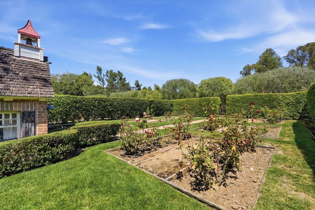 This serene garden scene features a series of raised planting beds filled with rose bushes, set against a backdrop of manicured hedges. To the left, a charming brick structure with a shingled roof and a small cupola adds a touch of classic architectural character to the landscape. The perspective is a wide-angle shot from the lawn, capturing the lush greenery and the peaceful, well-maintained outdoor space under a bright blue sky.