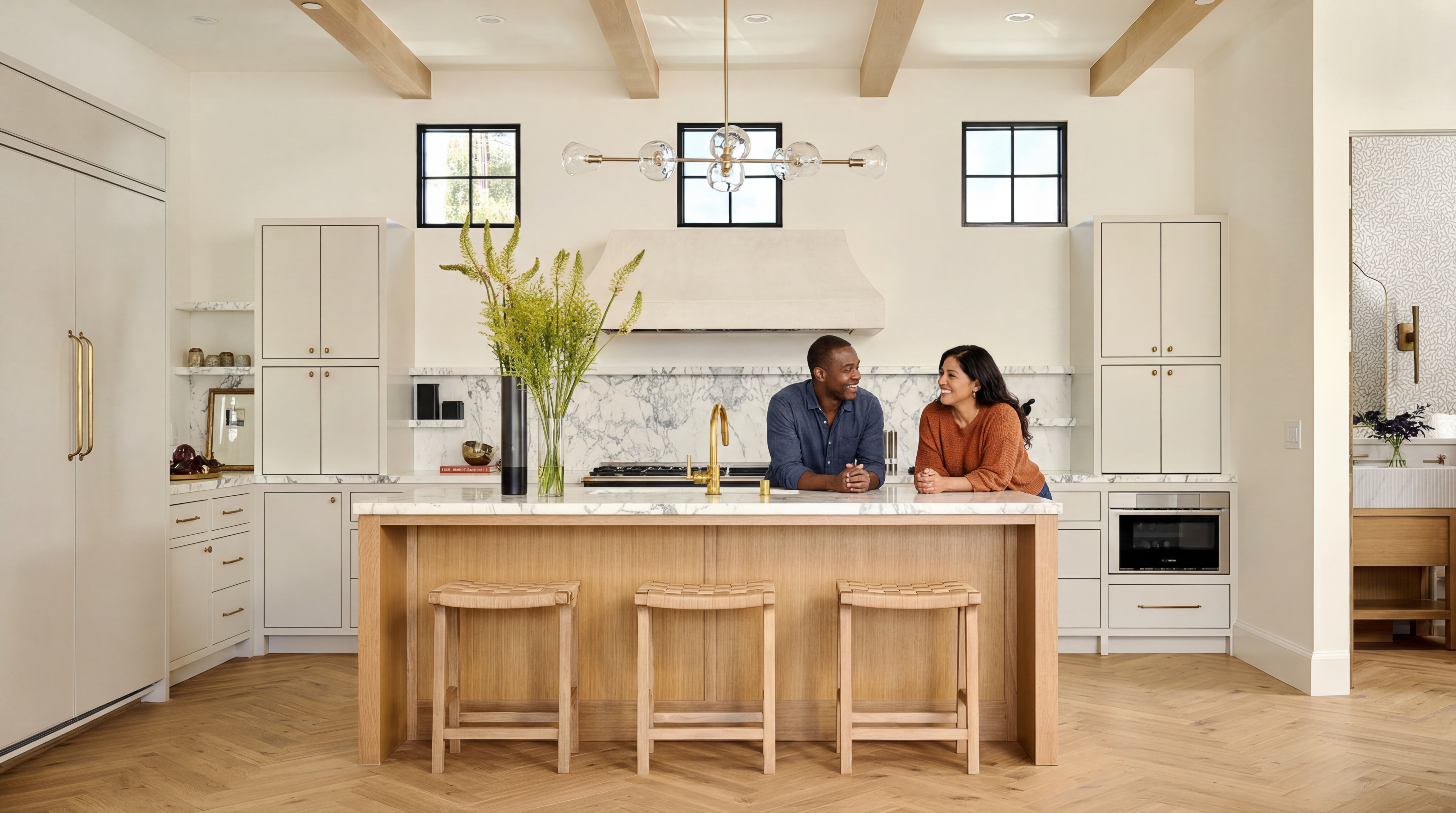 This stunning contemporary kitchen features light oak cabinetry, a large marble-topped island with seating, and elegant brass hardware. The space is anchored by a sophisticated marble backsplash and range hood, complemented by chic modern light fixtures and herringbone-patterned hardwood floors. A couple standing at the island adds a welcoming, lifestyle-focused aesthetic to this bright, high-end culinary space.